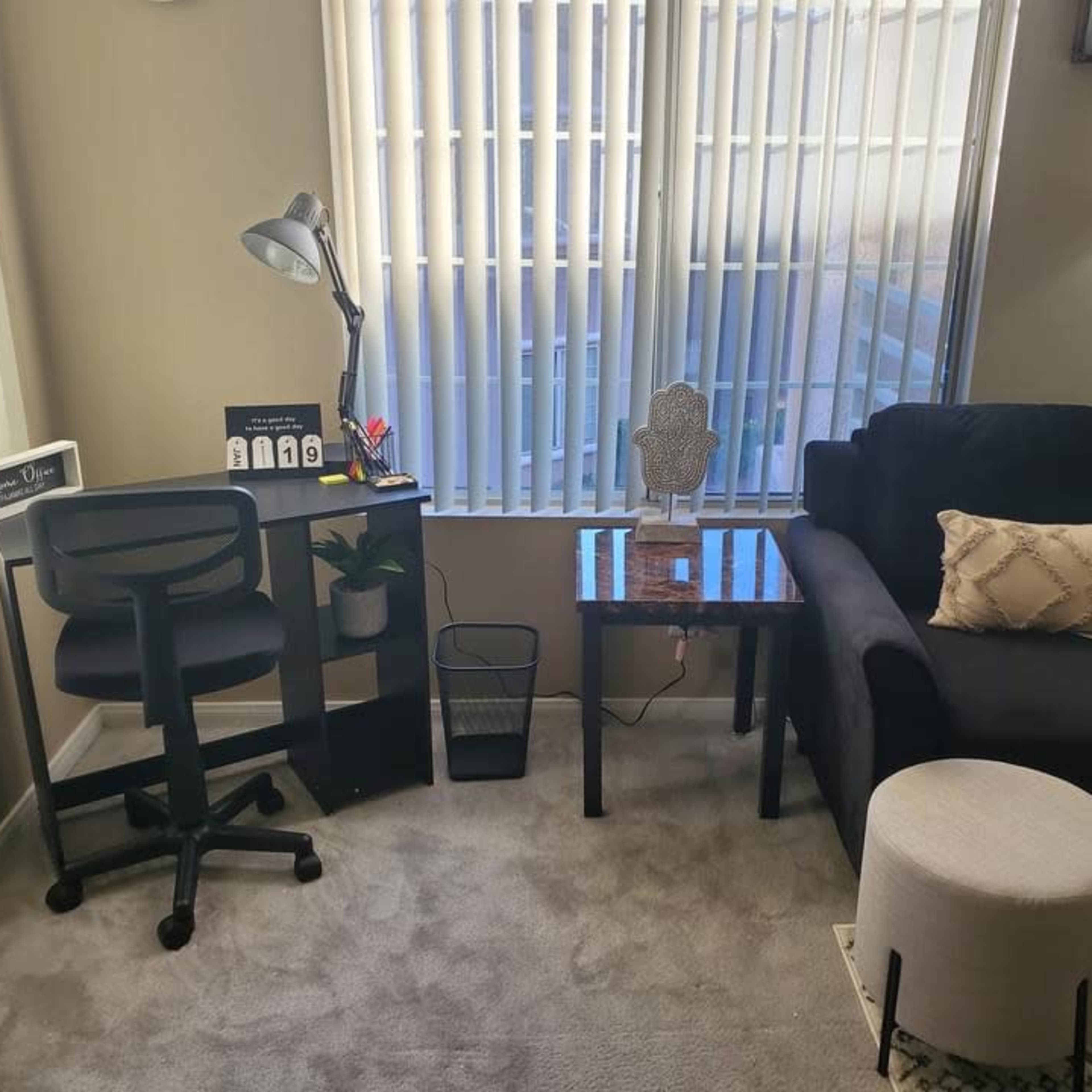The living space features a black chair, a small table with a decorative item, a white ottoman, a desk with a lamp, and large windows letting in natural light.