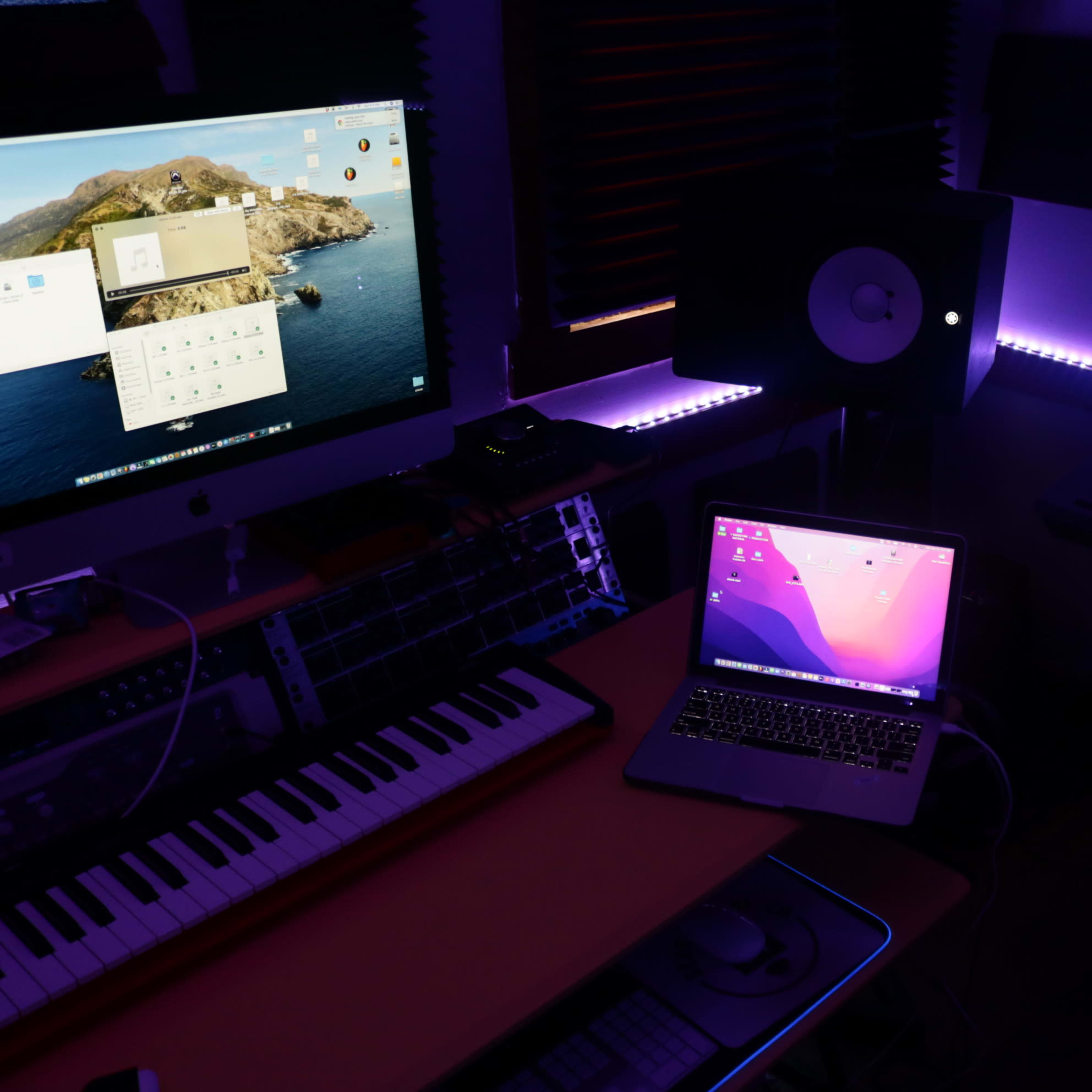 A music production studio with a desktop monitor, a laptop, keyboards, and ambient lighting.