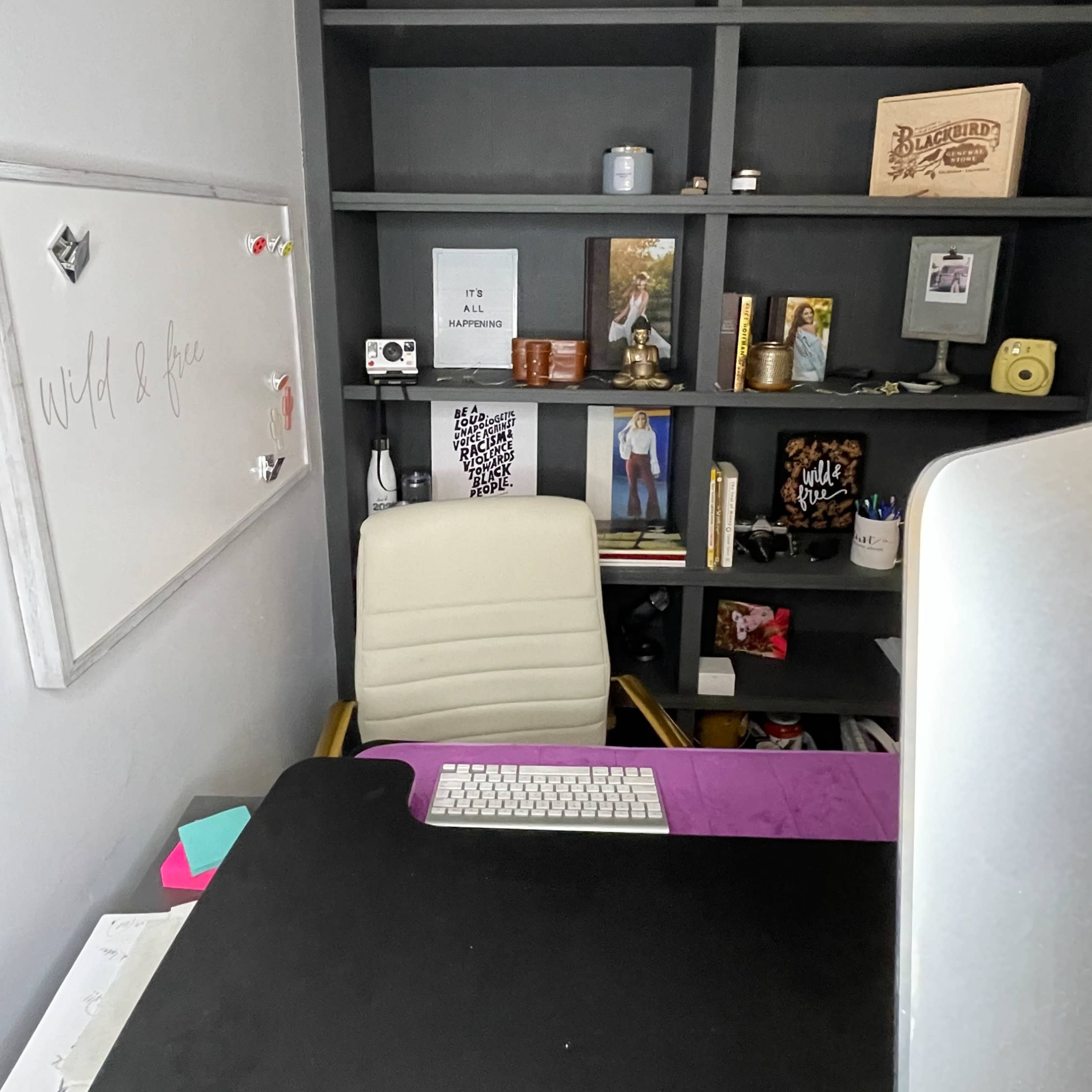 A modern office space with a white desk chair facing a computer monitor, surrounded by black shelves filled with various decorative items and photographs.