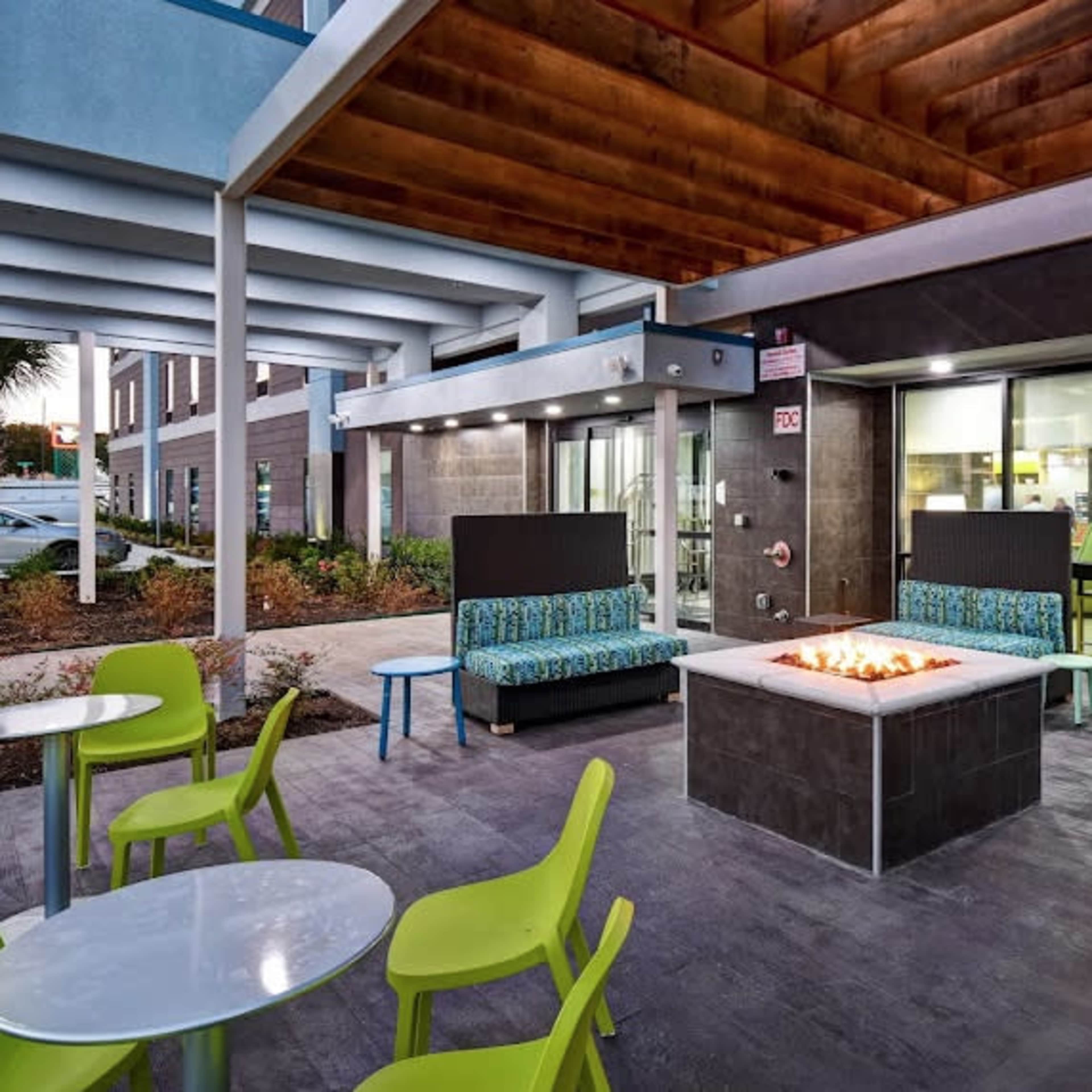 An outdoor seating area with colorful chairs and patterned couches surrounding a central fire pit, adjacent to a modern building.