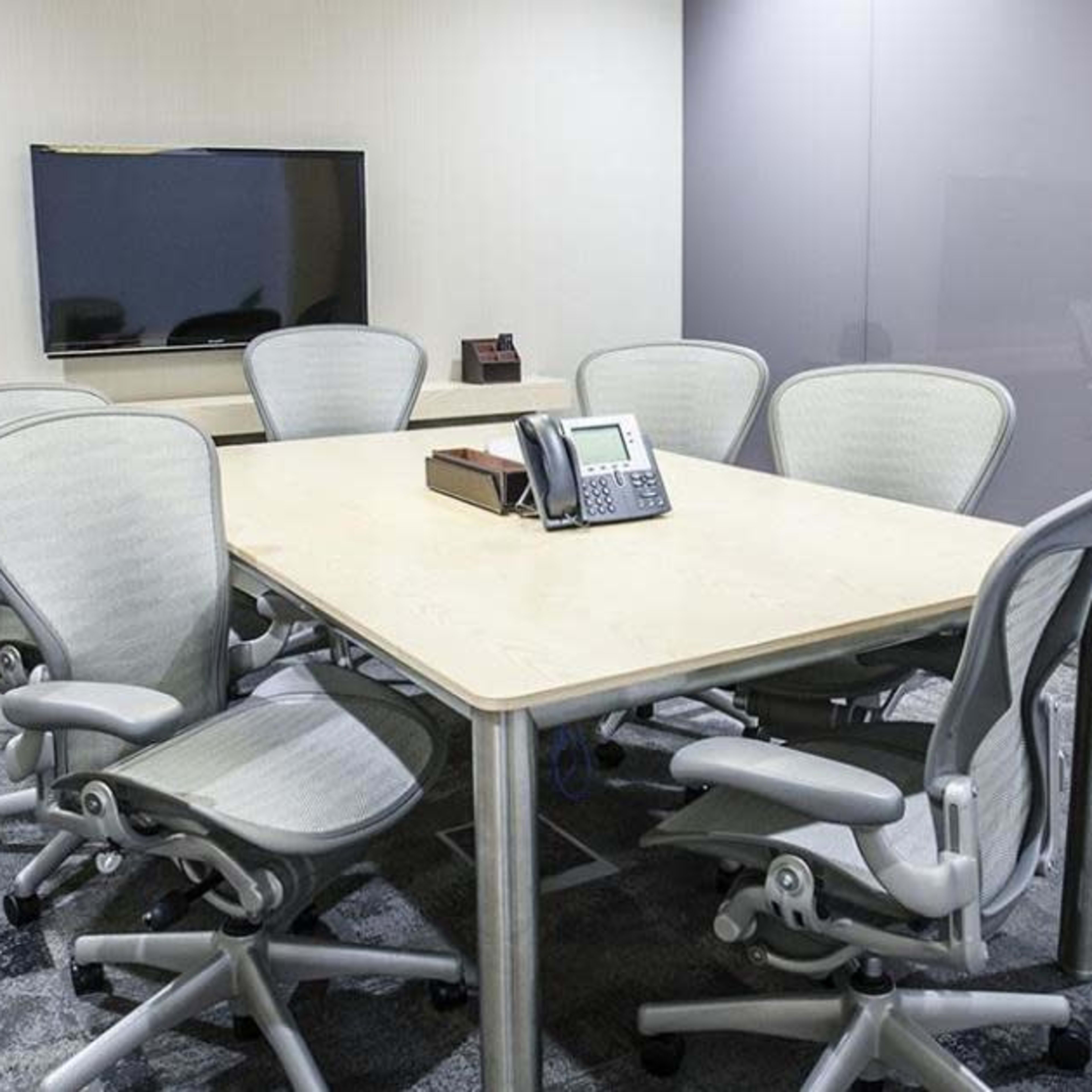 A conference room features a rectangular table surrounded by six ergonomic chairs, with a television on the wall and a phone at the center of the table.