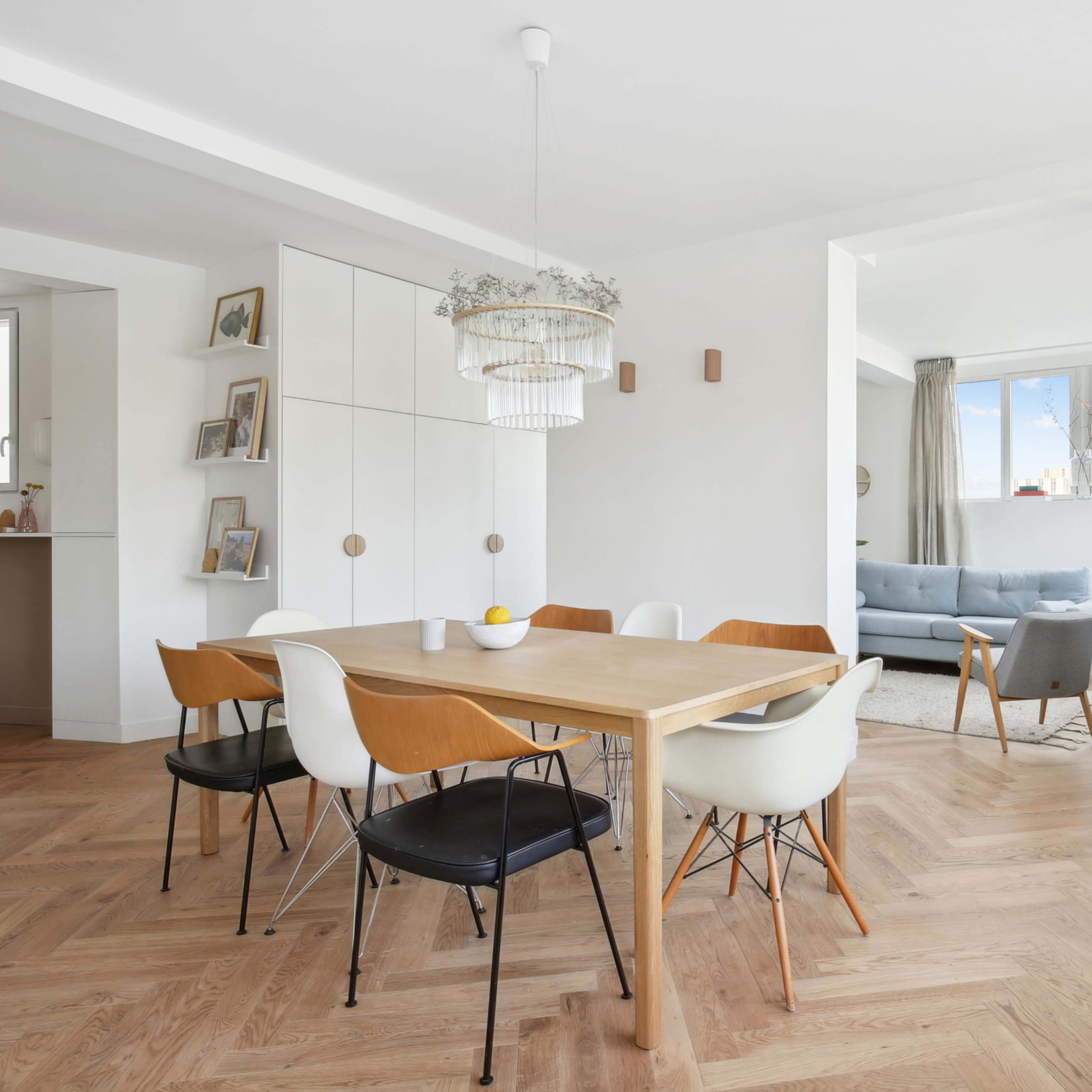 A dining area with a wooden table surrounded by chairs, adjacent to a living room featuring a sofa and large windows.