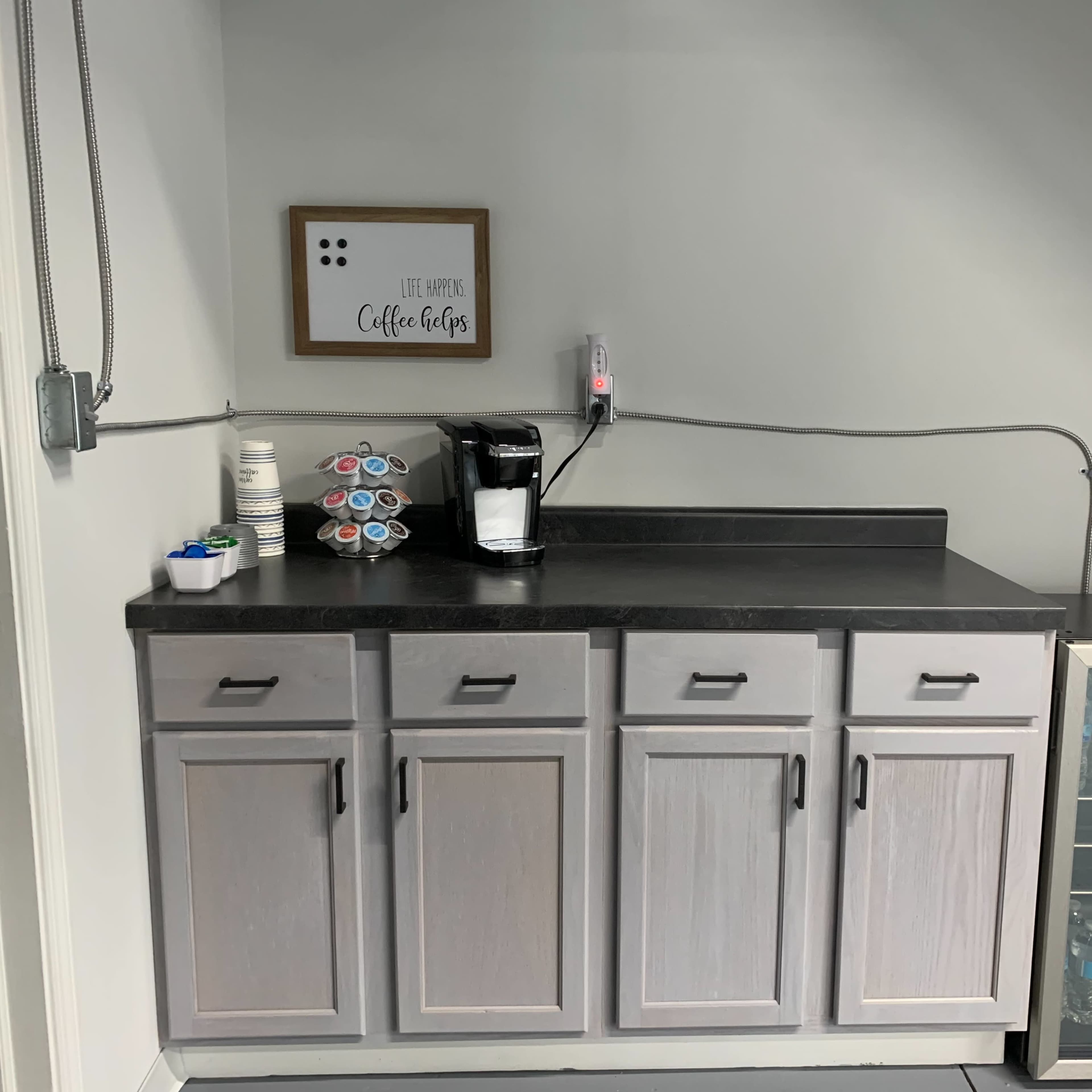 A small coffee station with a coffee maker, stacked cups, and a mini refrigerator filled with bottled water.