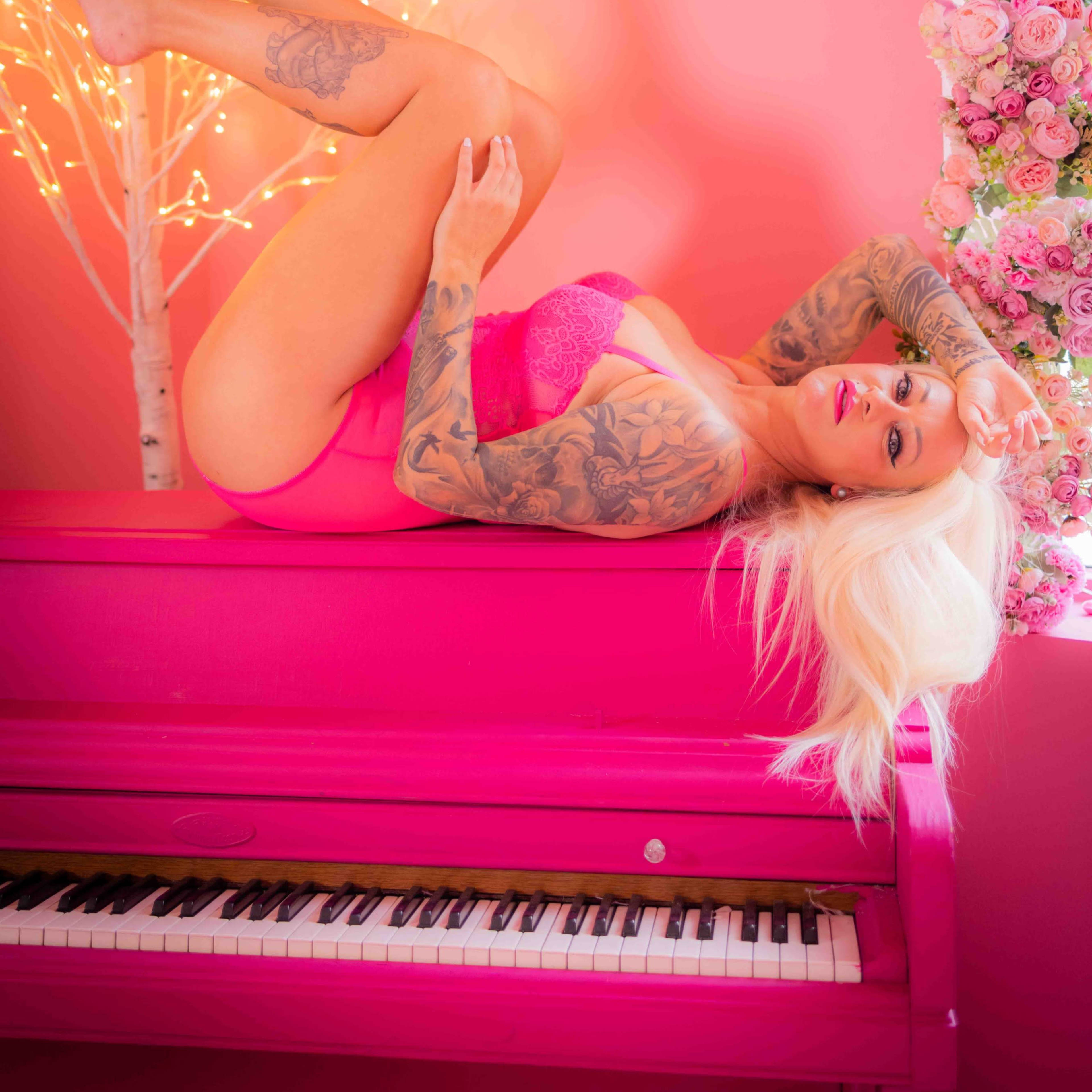 A person with long blonde hair and tattoos poses on a pink piano against a wall decorated with flowers and fairy lights.