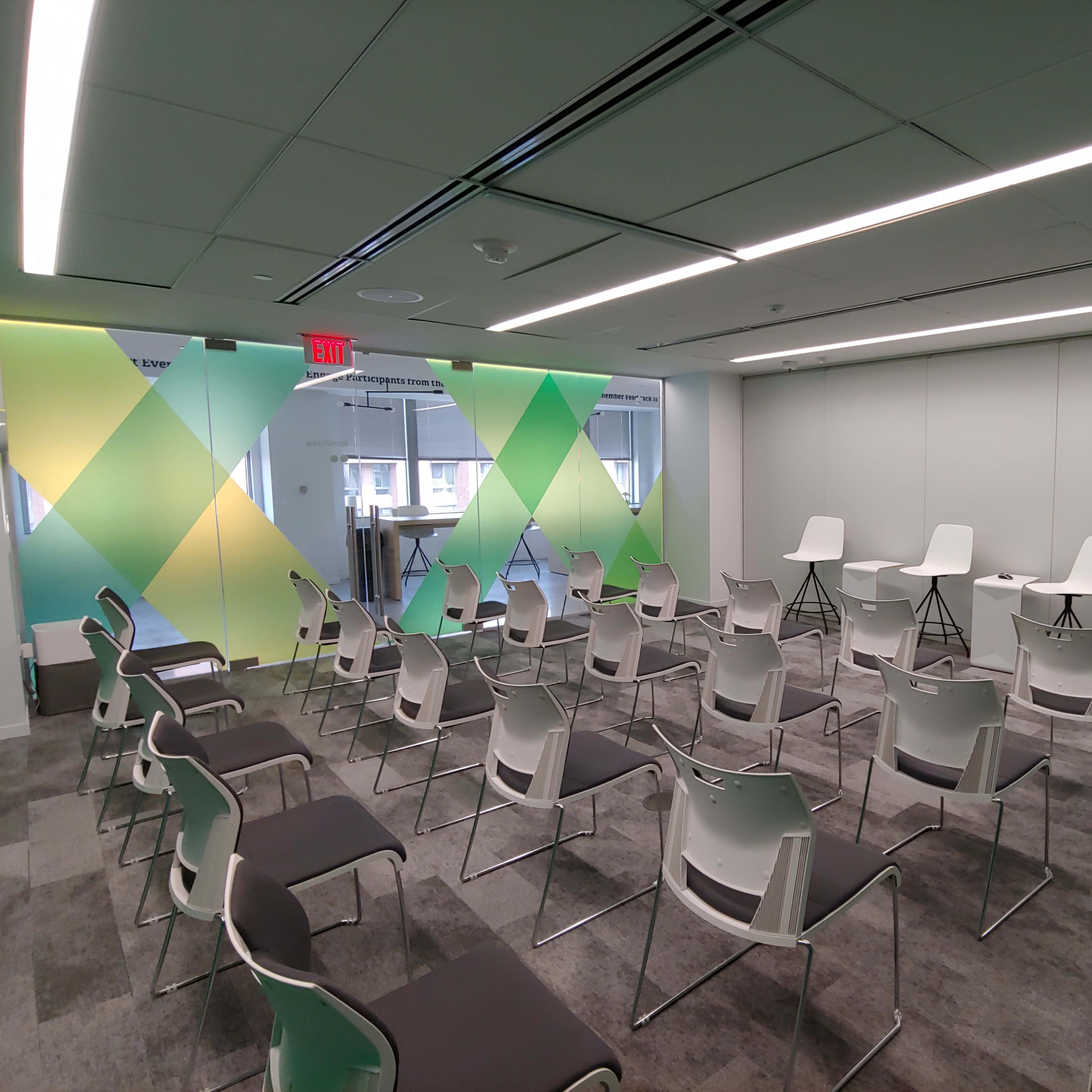 A meeting room with neatly arranged chairs facing a small stage area, featuring colorful wall graphics and large windows.