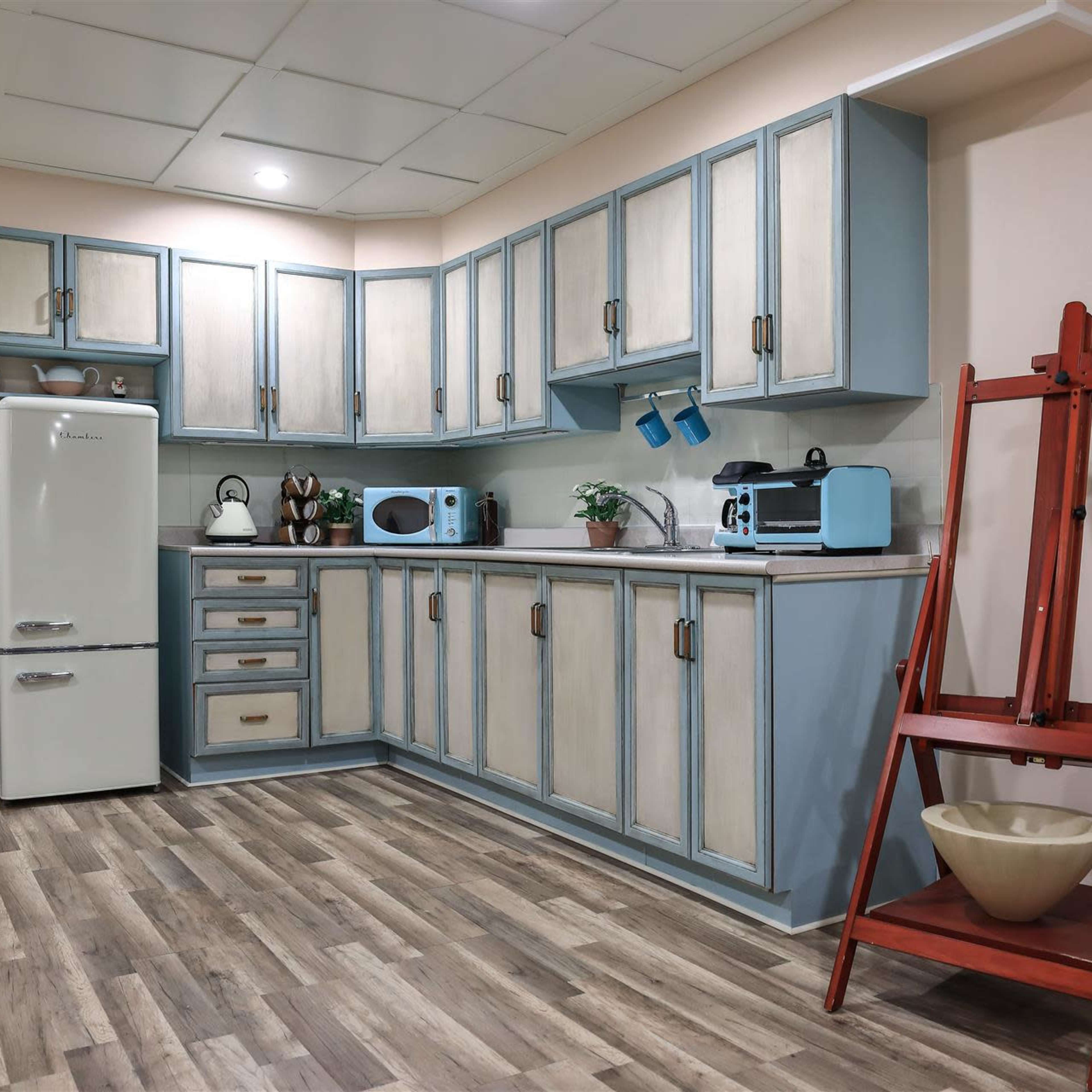 A modern kitchen with light blue and cream cabinets, a vintage-style refrigerator, a few small kitchen appliances, and a red wooden easel with a bowl on a stand.