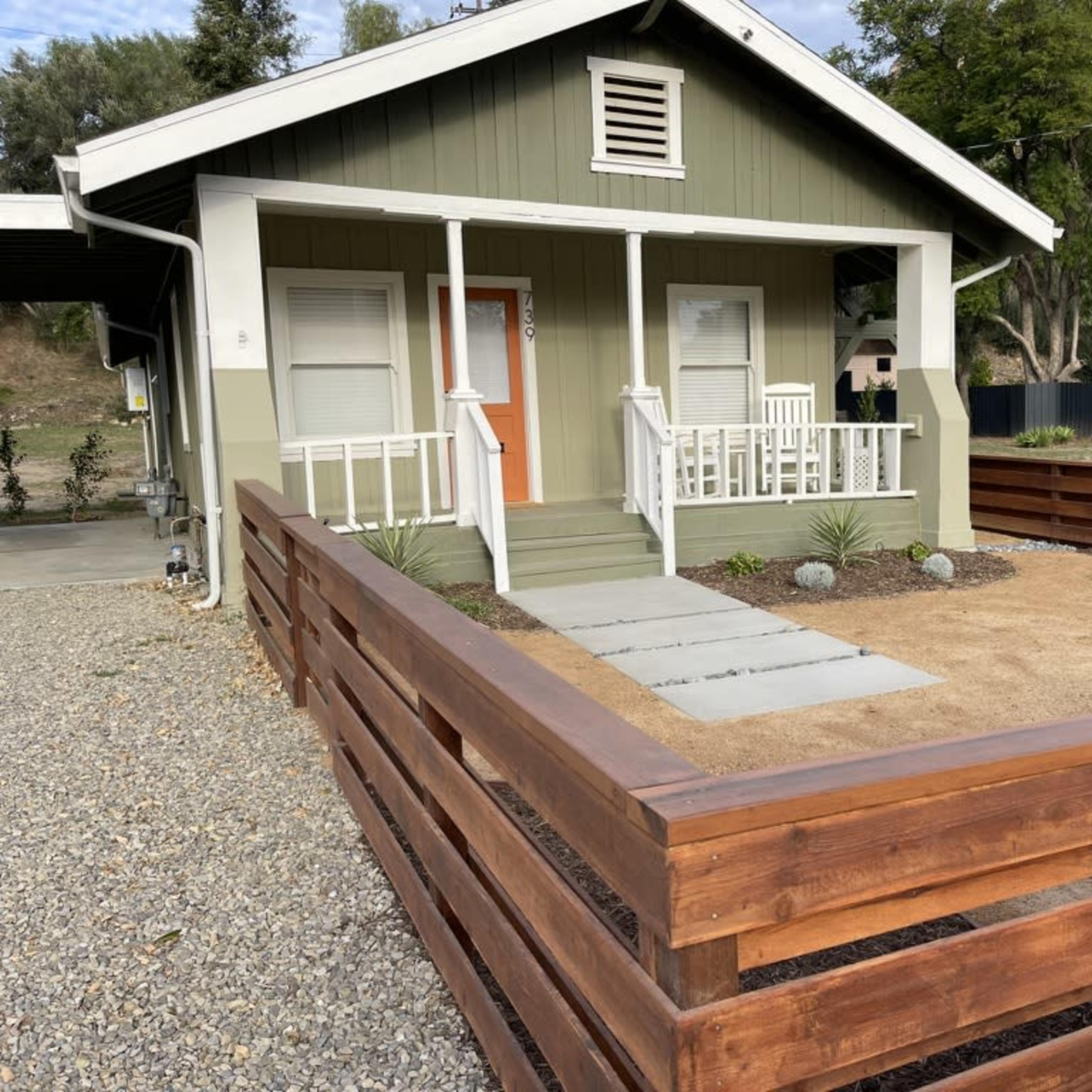 A small house with a green exterior, a front porch, and a fenced yard featuring gravel and sparse landscaping.