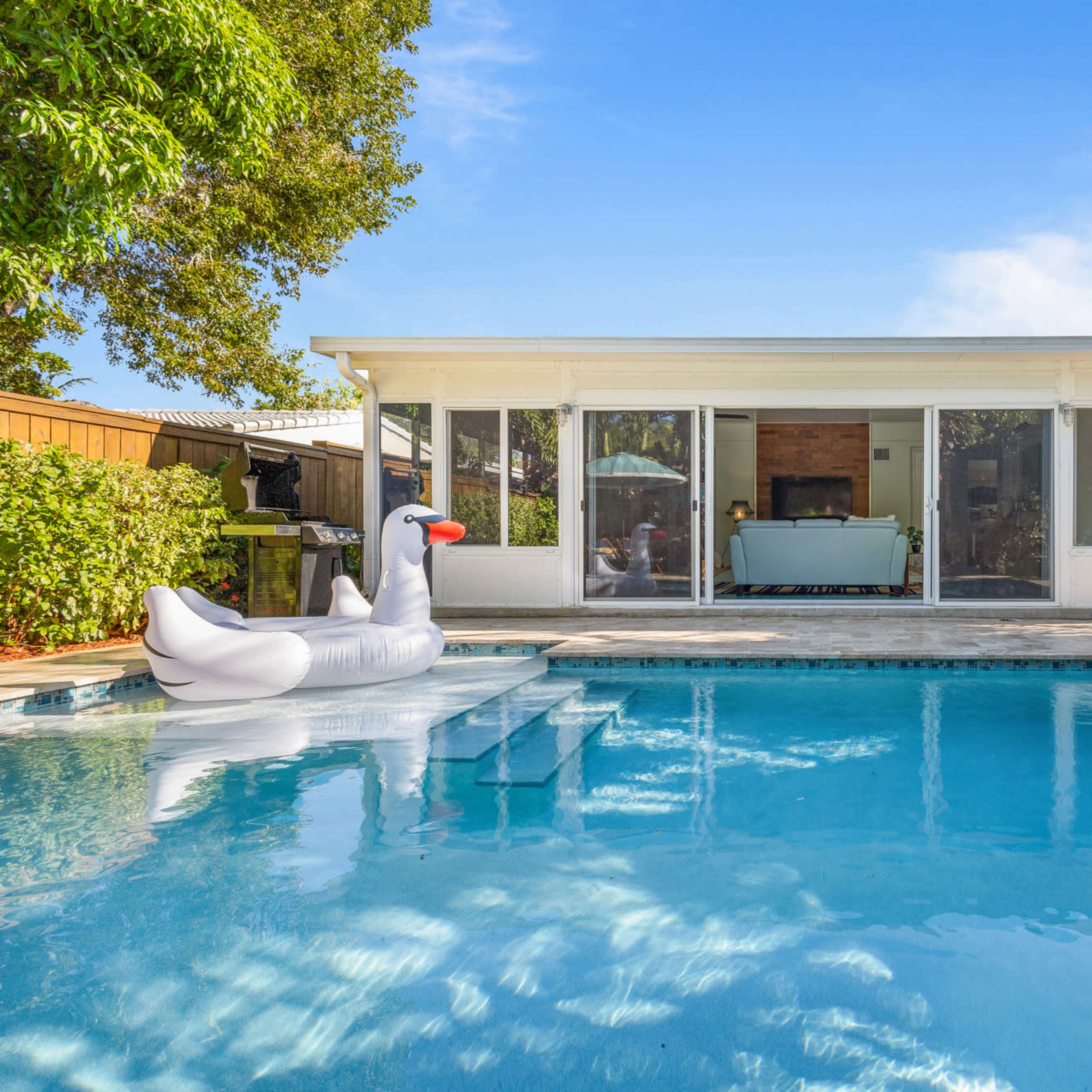 A swimming pool with a swan-shaped float is located in front of a modern house surrounded by palm trees.