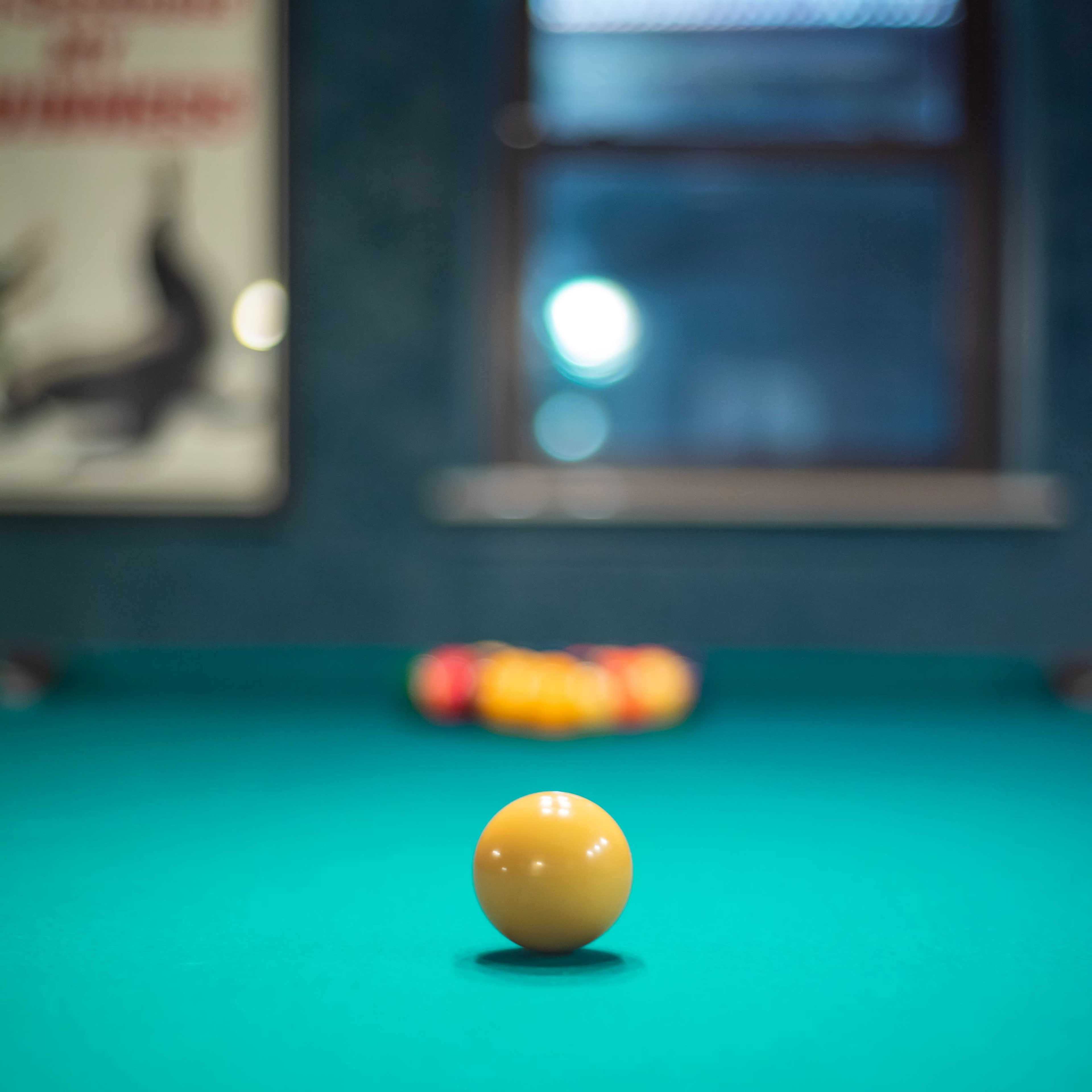 A single yellow billiard ball rests on a green felt pool table, with a blurred rack of colored balls in the background.