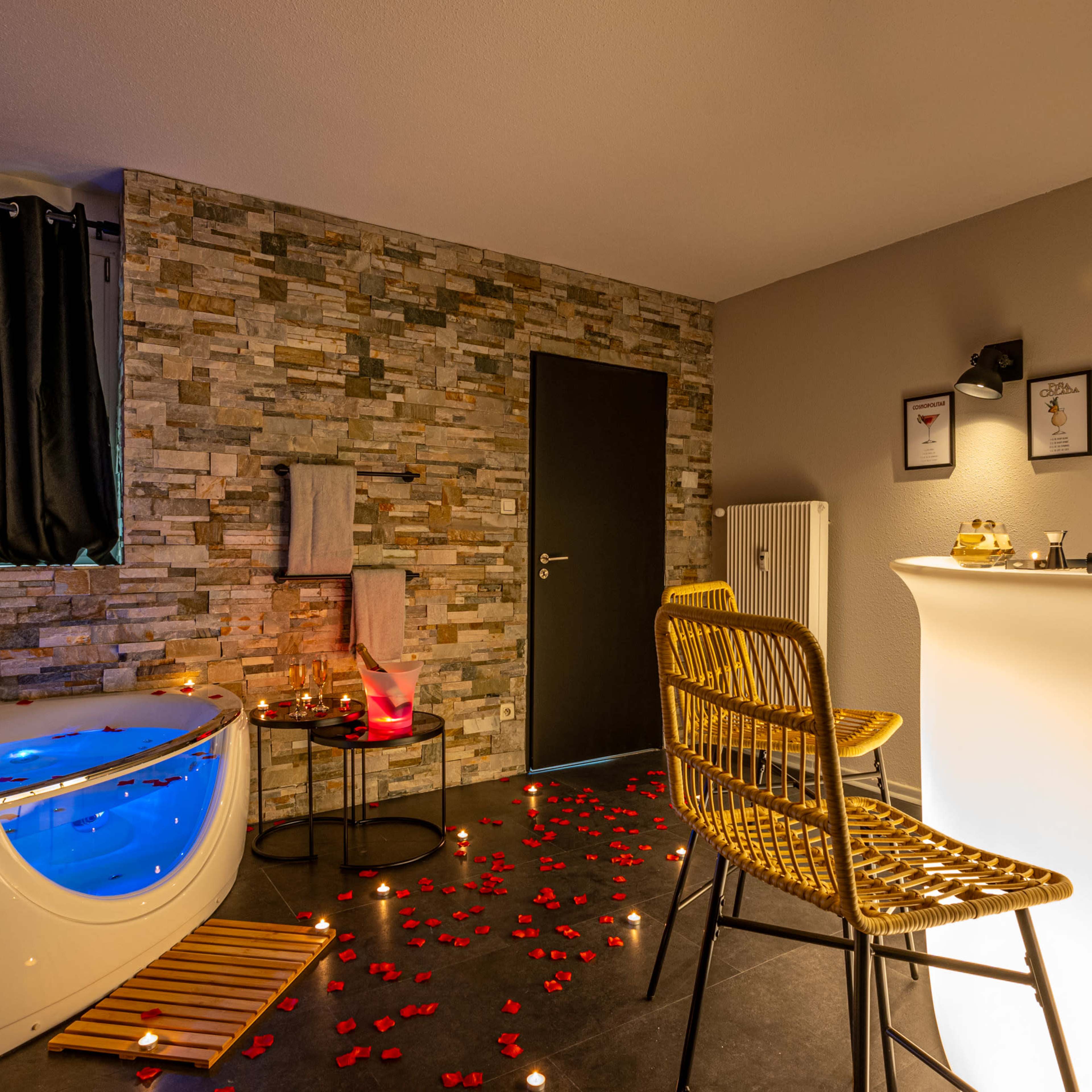 A modern bathroom with a freestanding tub, decorative stone walls, and a bar area featuring a chair and a small table adorned with rose petals and candles.