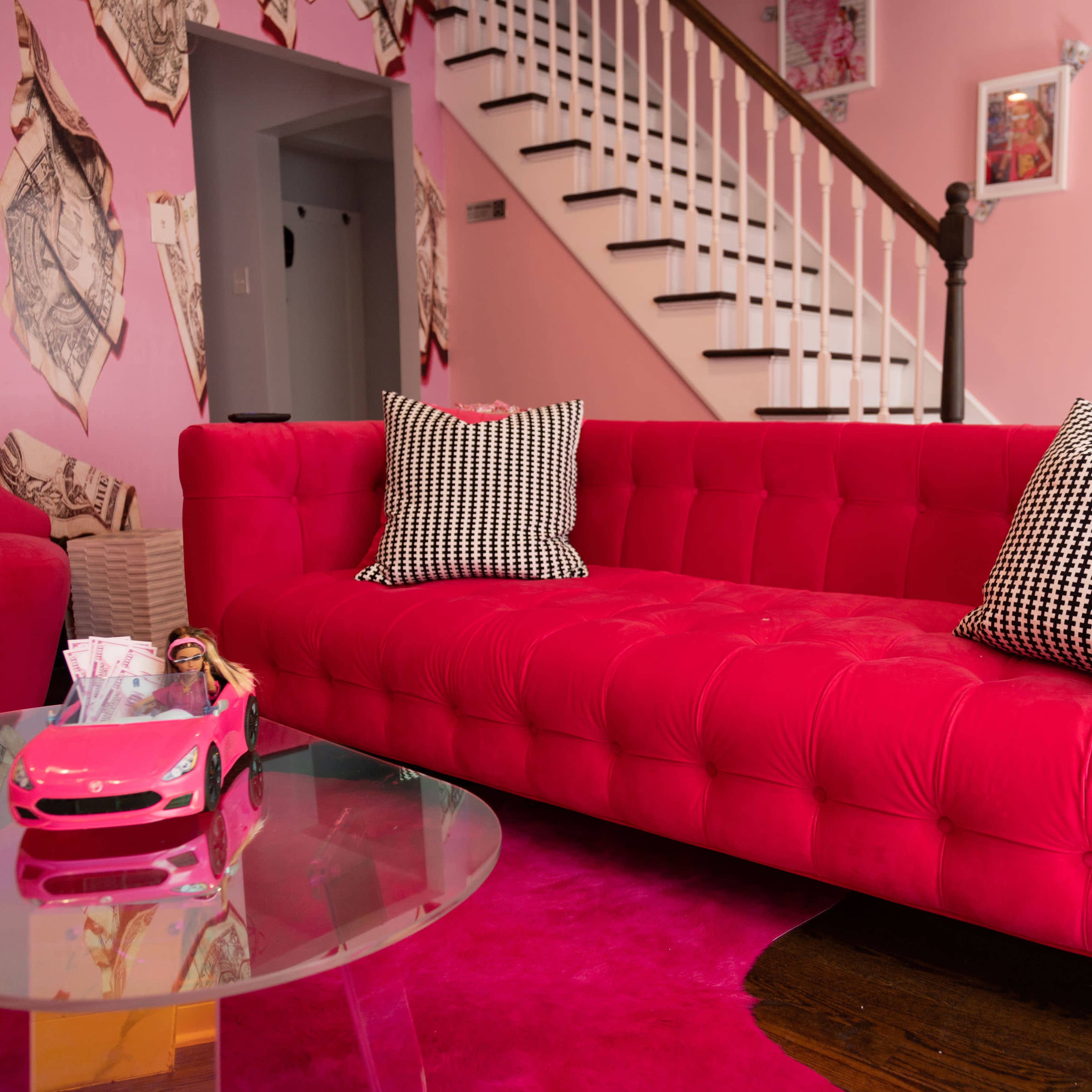 The scene features a bright pink velvet sofa with black and white checkered pillows, positioned next to a glass table and a toy car on a matching pink rug in a room decorated with pink walls and framed photos.