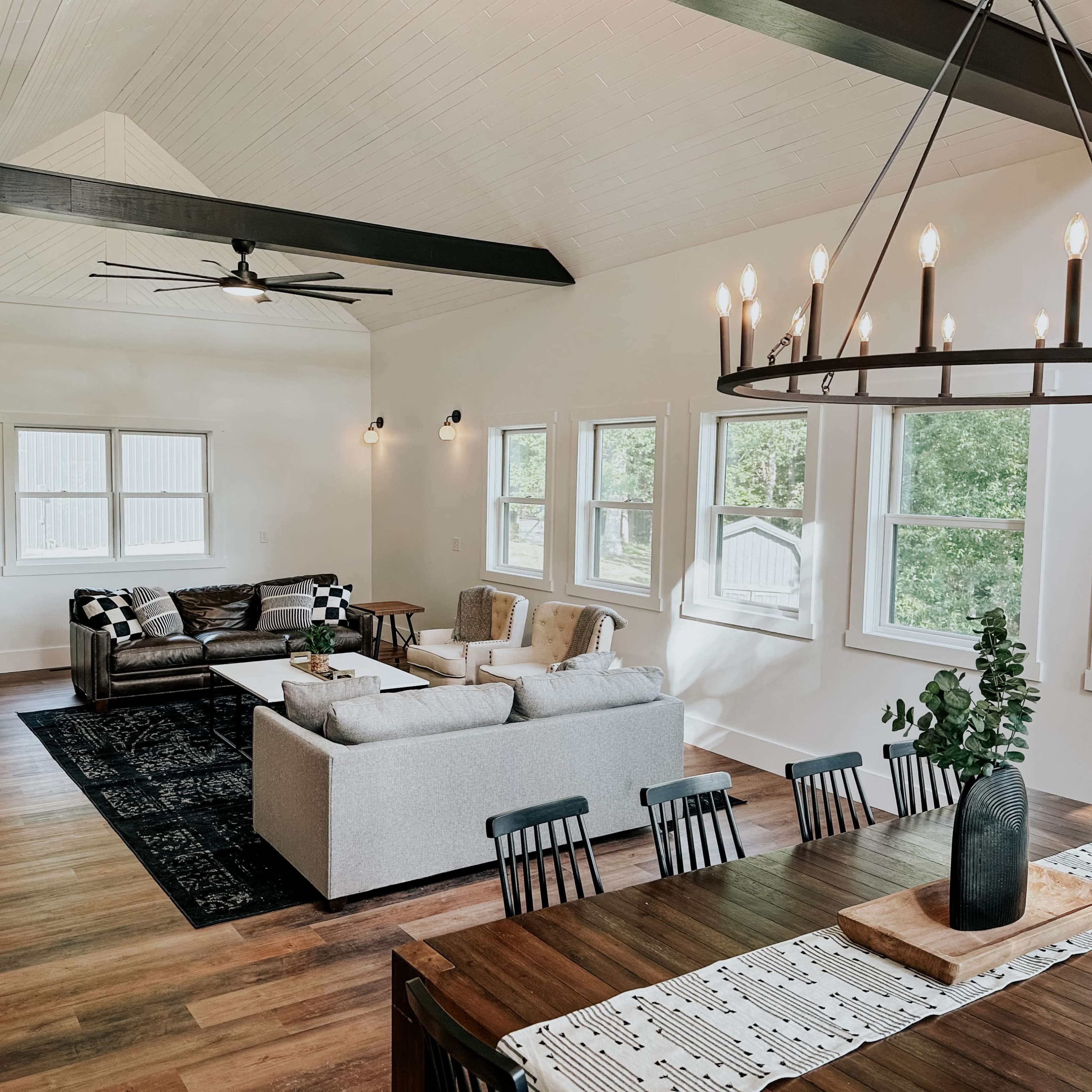 A spacious living area features a mix of modern furniture, large windows, and a dining table set in a well-lit room with wooden flooring.