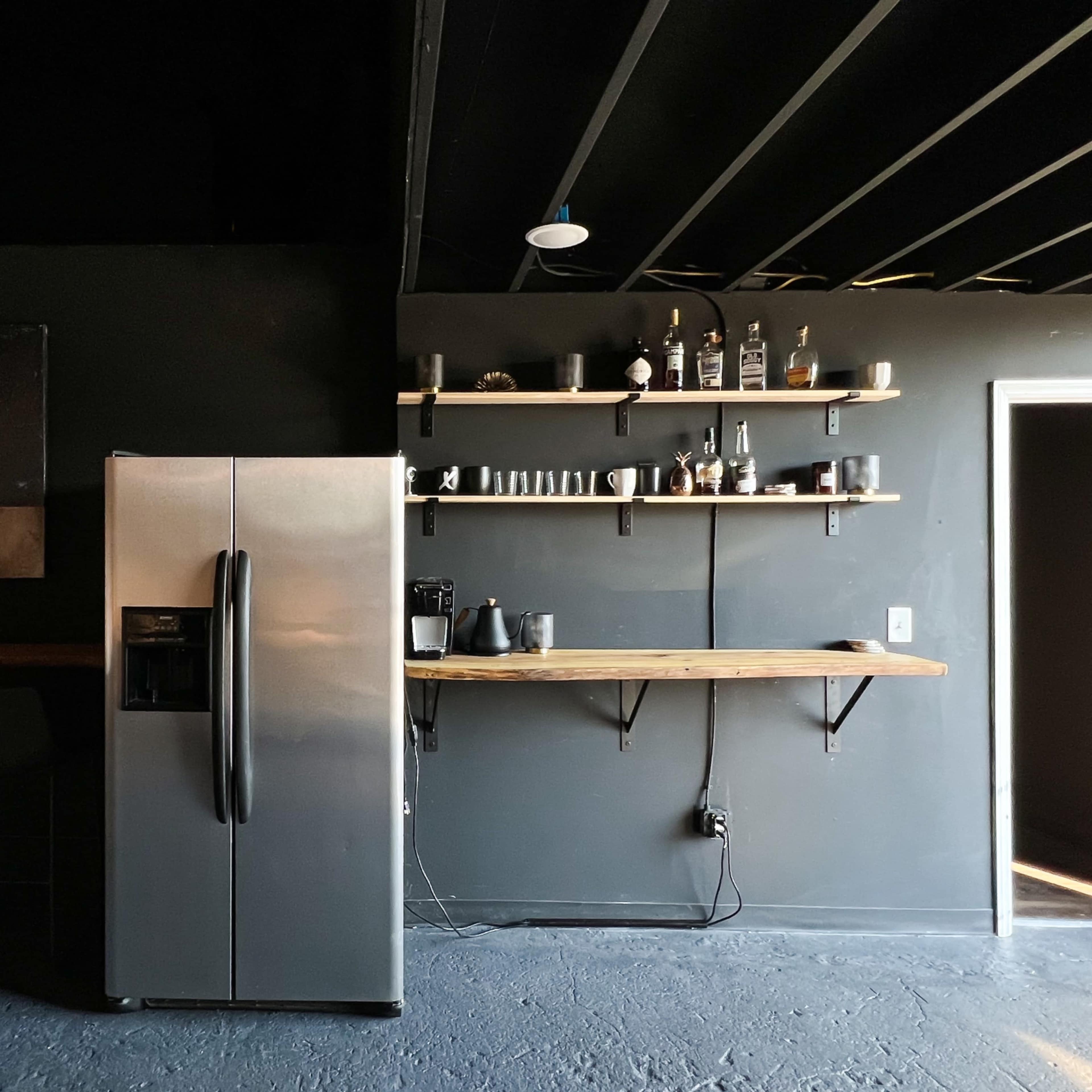 A modern kitchen area featuring a stainless steel refrigerator, a wooden bar counter with stools, and shelves displaying various glassware and appliances against a dark wall.