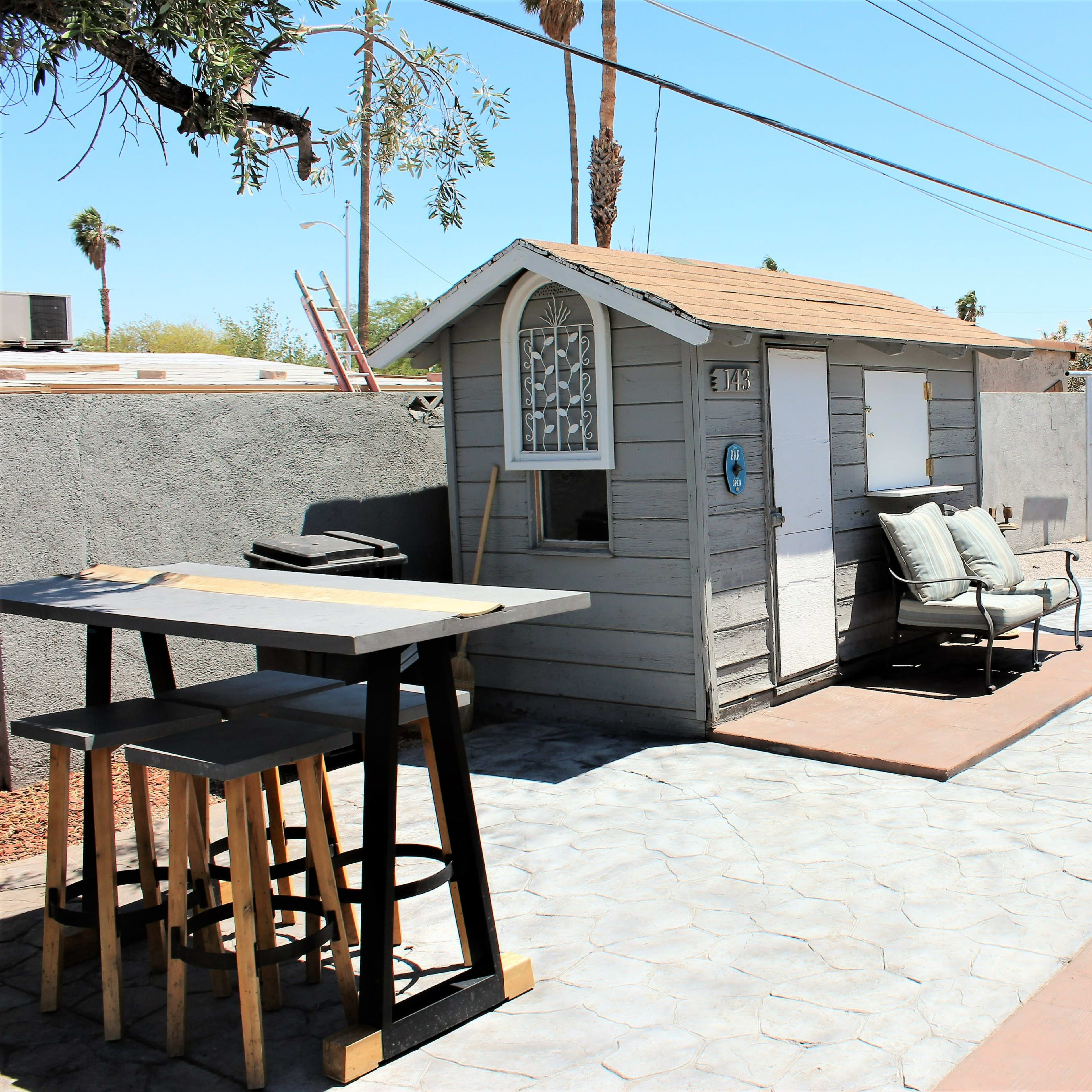 A backyard with a playhouse, a wooden tree sculpture, and a concrete patio area with a table and chairs under a clear blue sky.