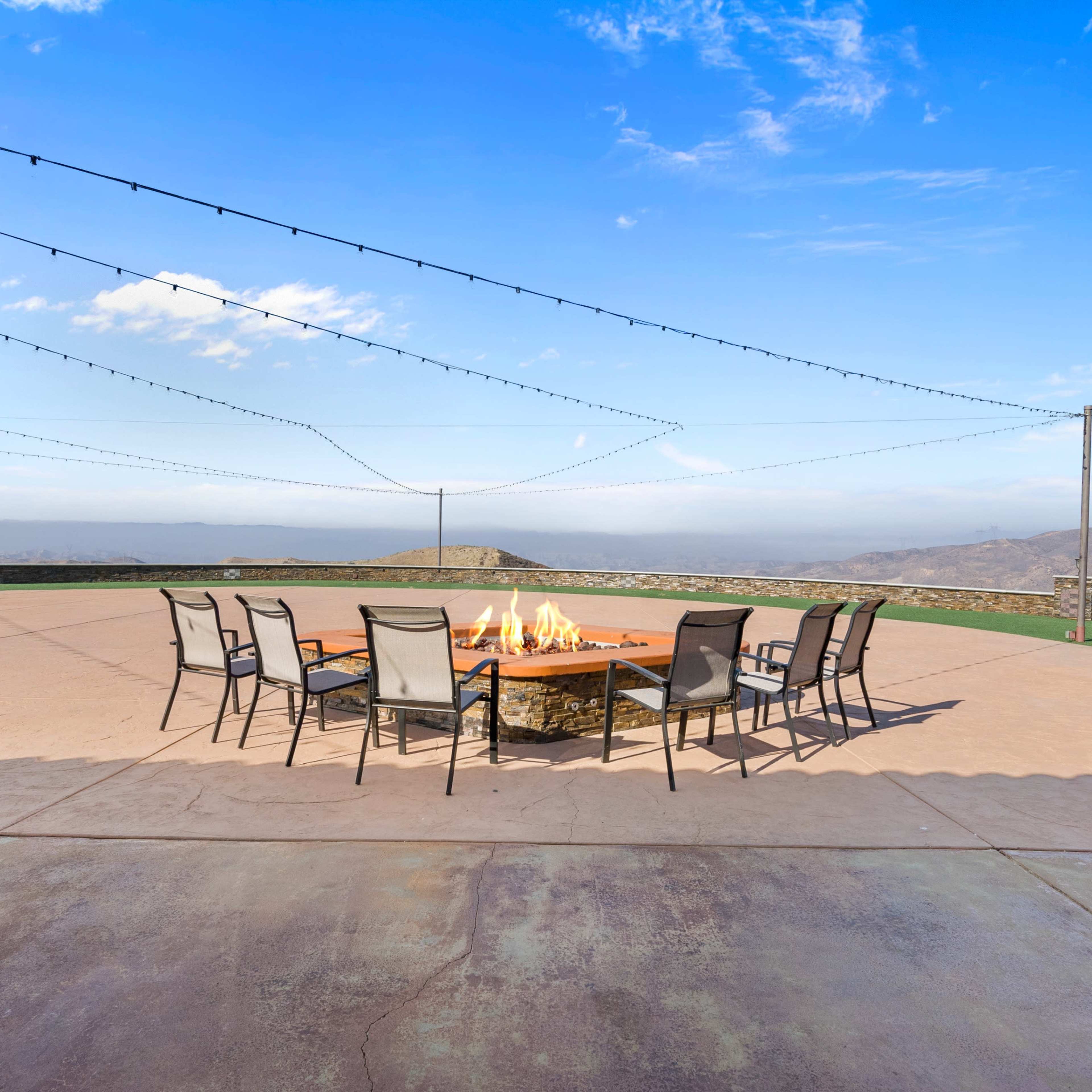 A circular fire pit surrounded by ten chairs is set on a concrete patio with mountains in the background under a clear sky.