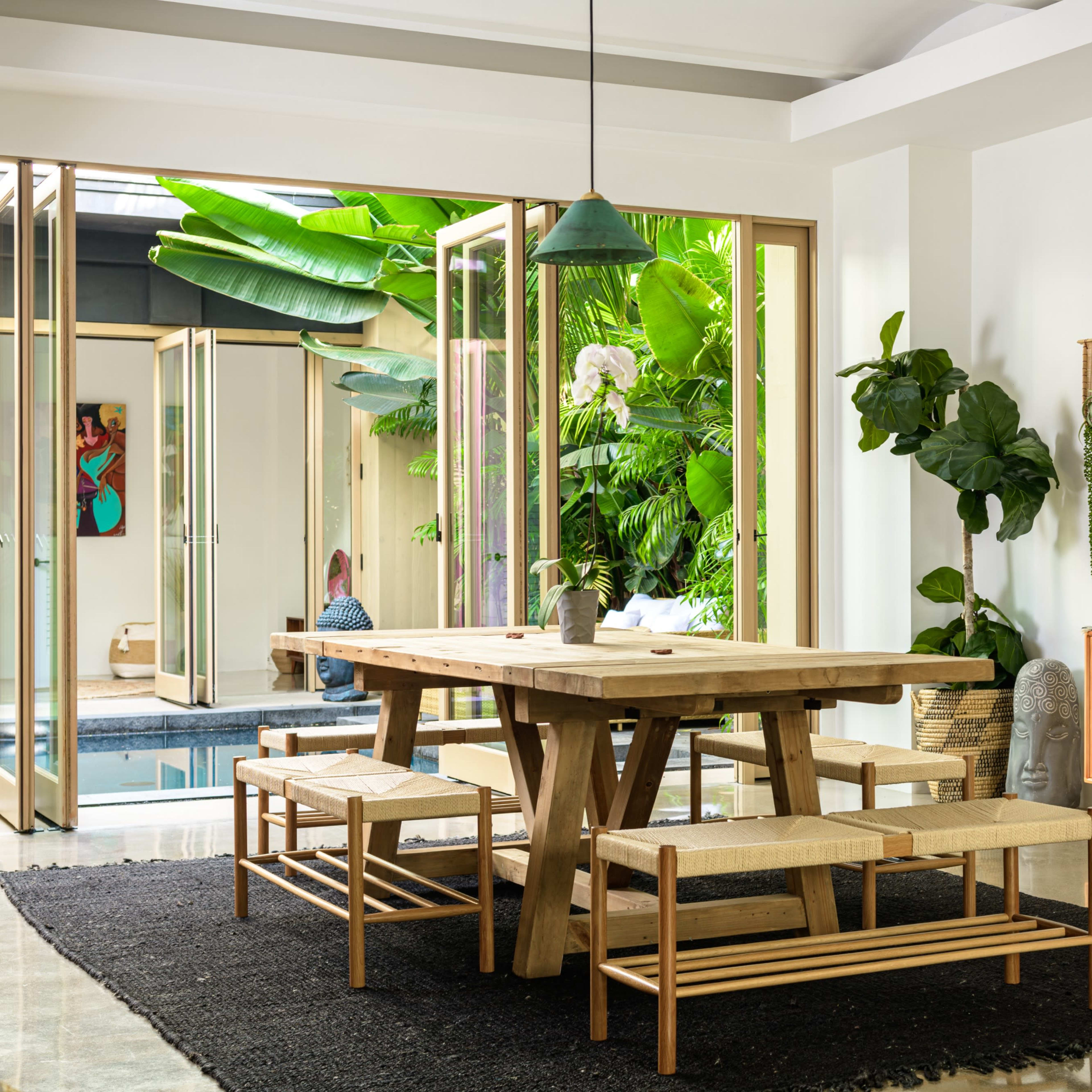 A dining area features a large wooden table with benches, surrounded by plants and opening to a poolside view through glass doors.