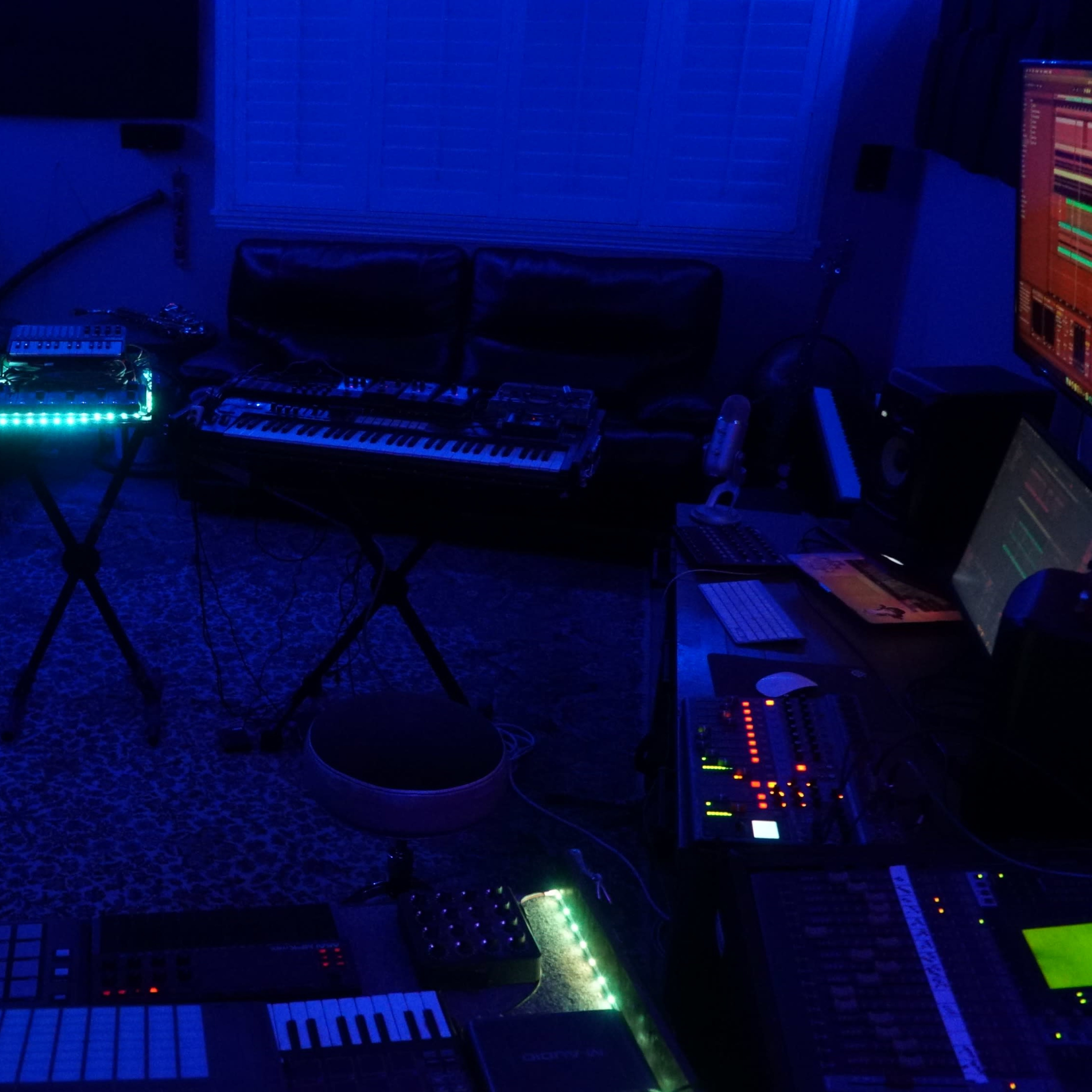 A dimly lit music studio features electronic instruments arranged on stands with soft blue lighting and a couch in the background.
