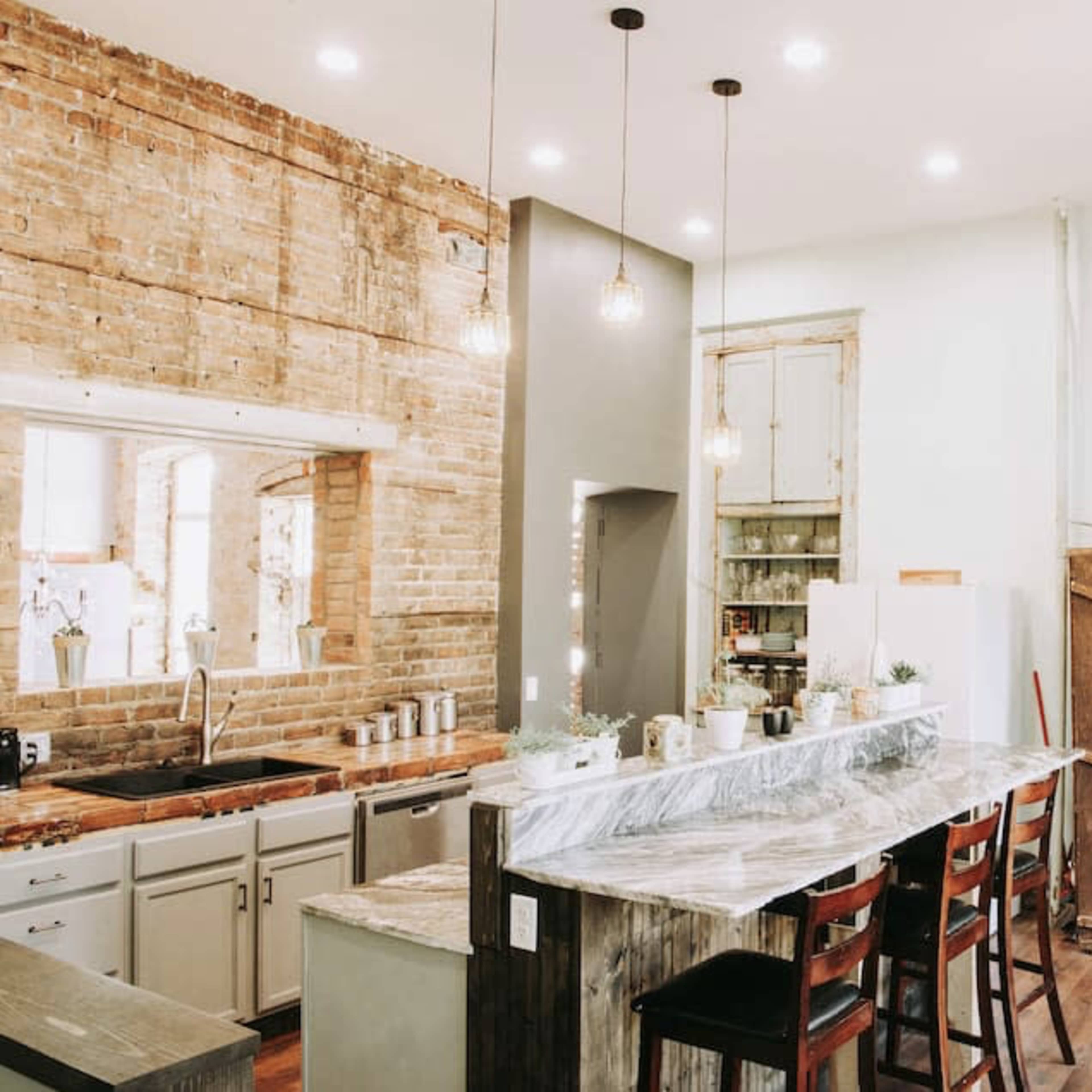 A modern kitchen with exposed brick walls, a large marble island, wooden bar stools, and open shelving displaying various kitchen items.