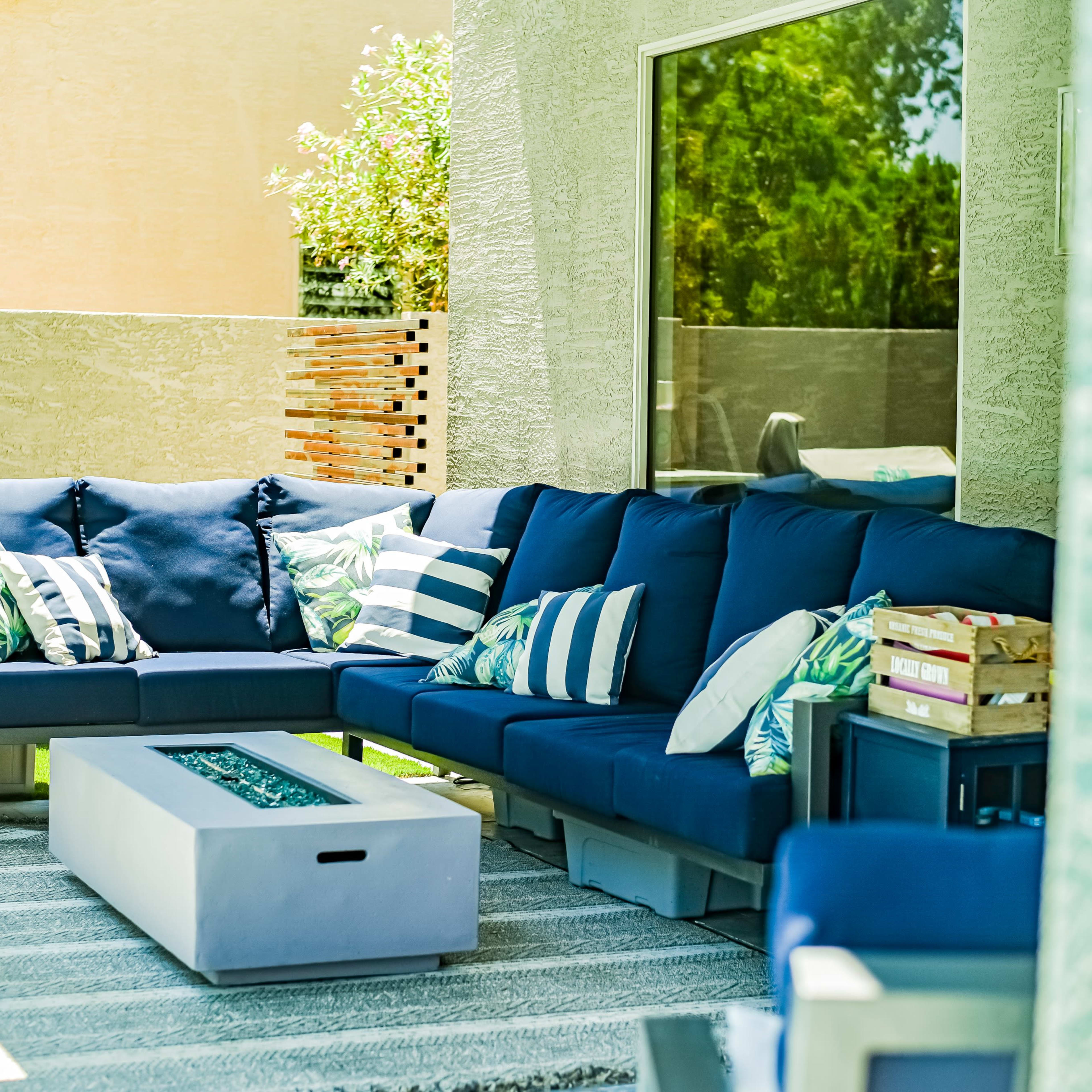 A modern outdoor seating area features a navy sectional sofa with decorative cushions, a rectangular fire table, and a textured rug.
