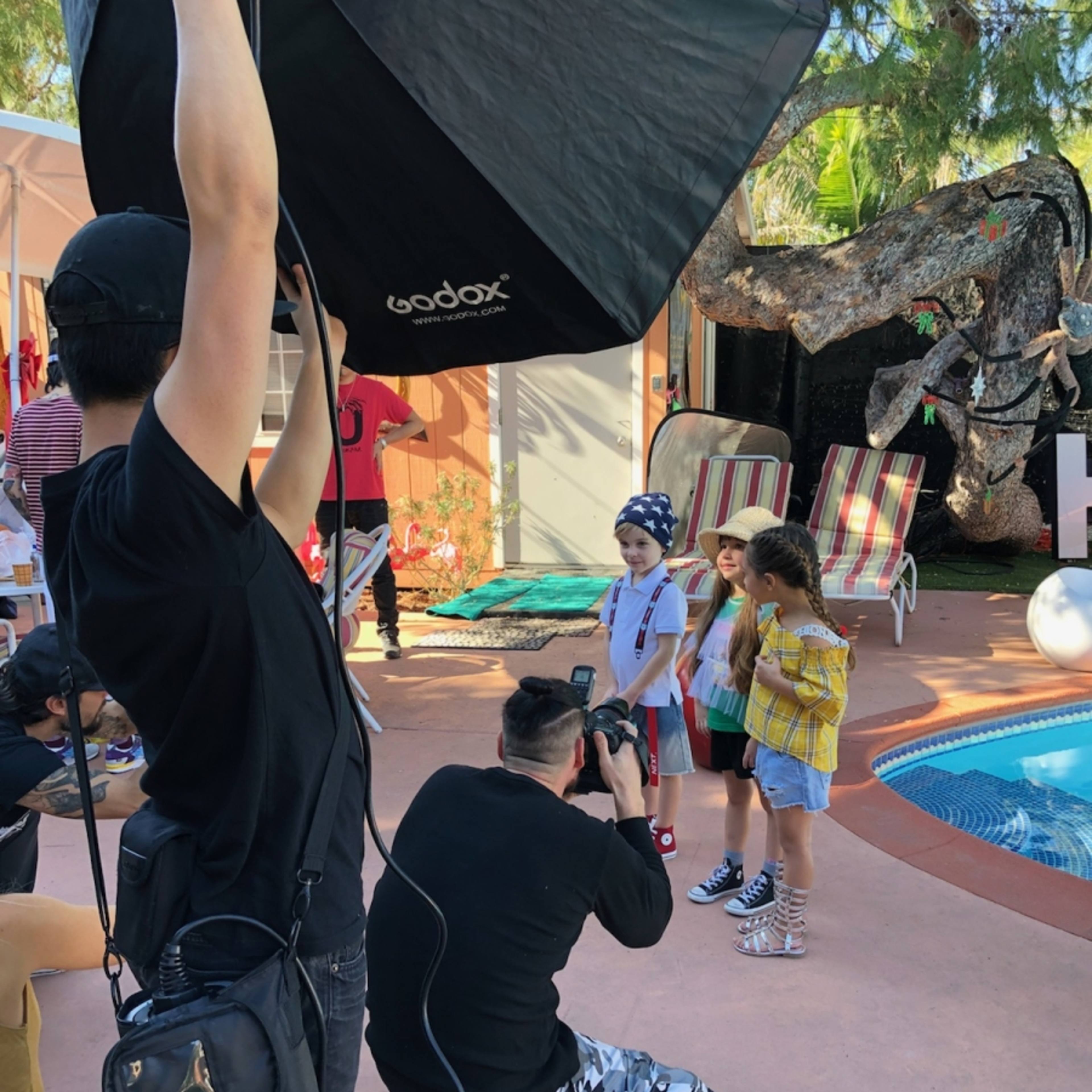 A photographer captures two children posing under a large softbox light near a pool surrounded by lounge chairs and greenery.