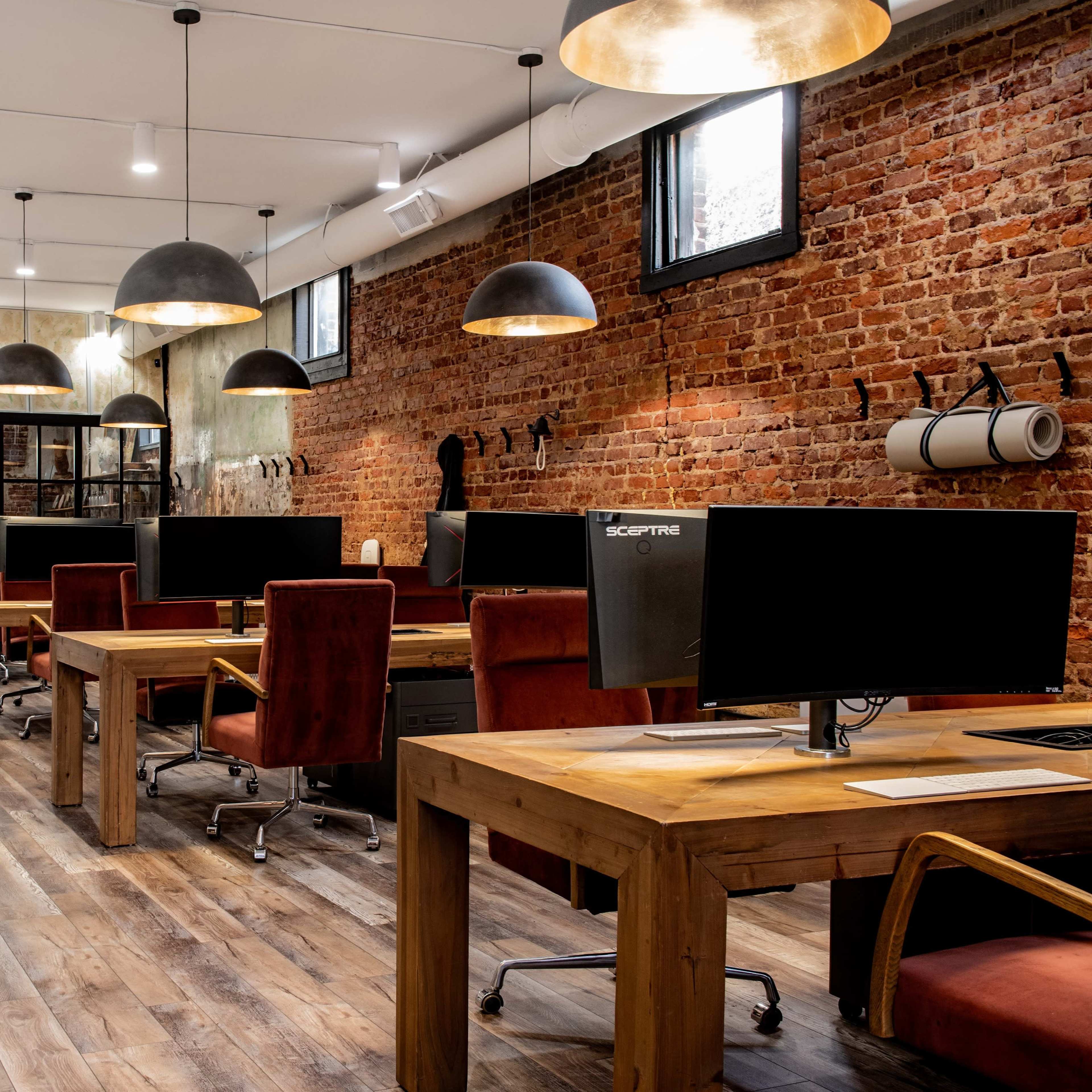 A modern office space with wooden desks, large computer monitors, and an exposed brick wall, illuminated by pendant lights.