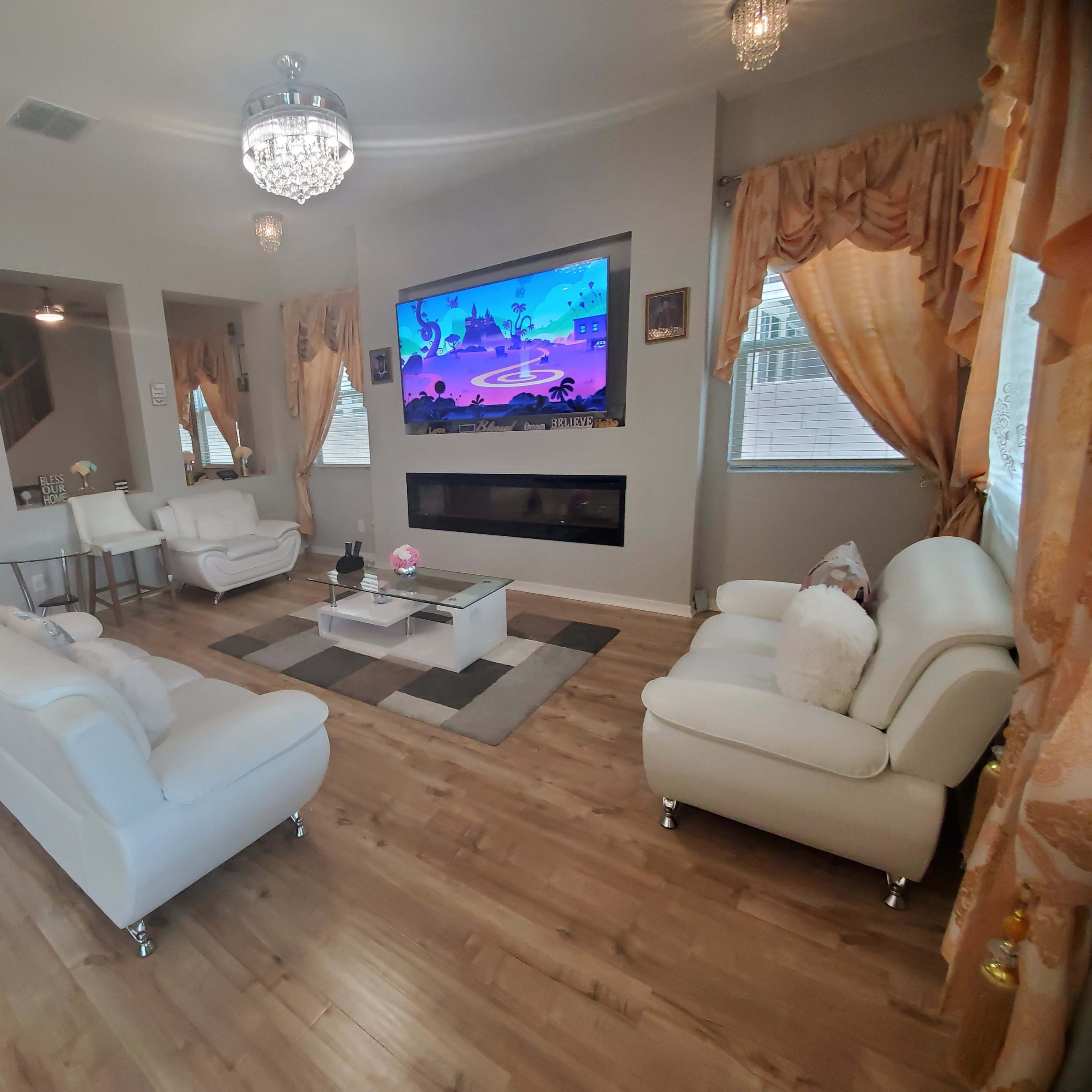 The living room features two white sofas, a glass coffee table, and a large wall-mounted TV, with decorative curtains and a modern fireplace.