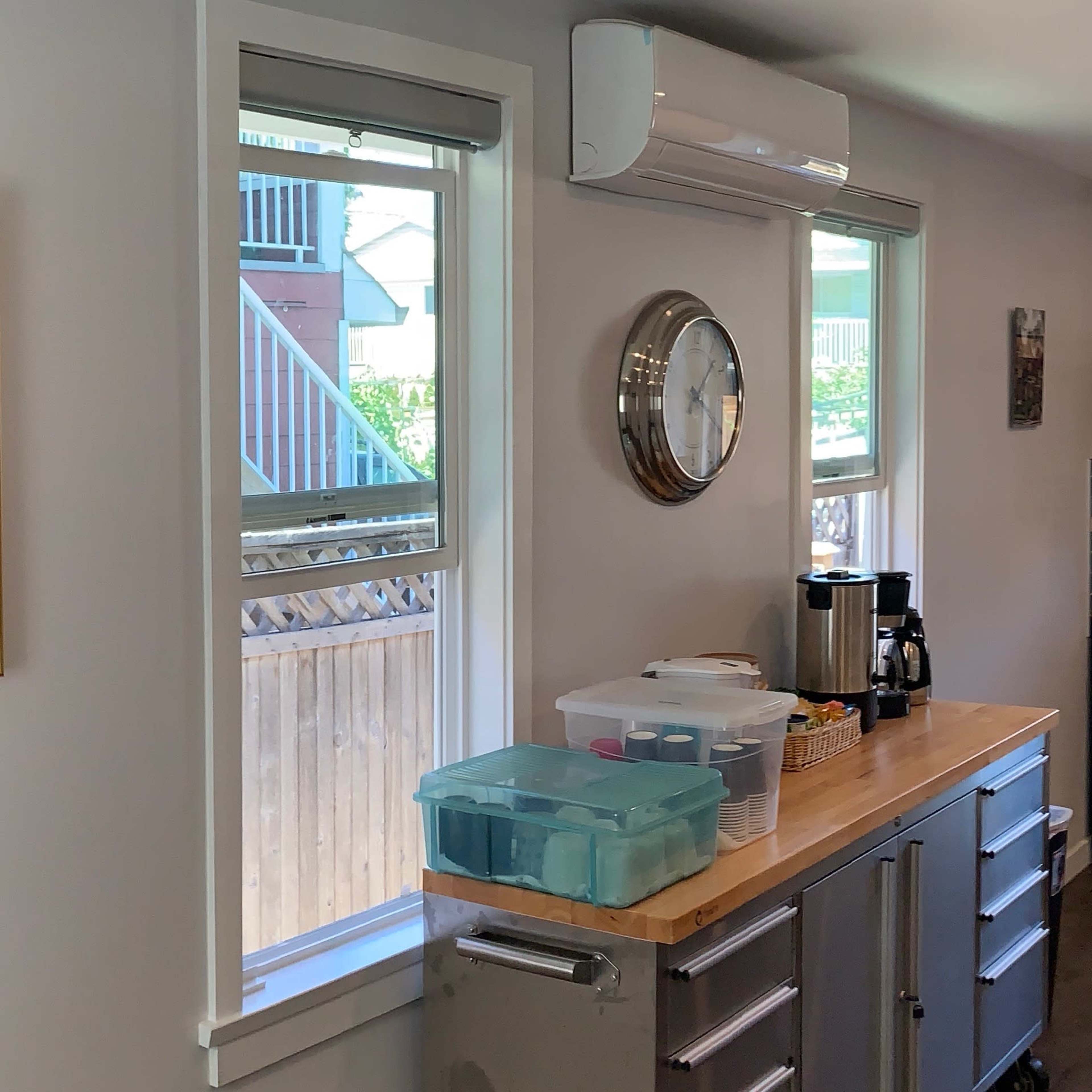 A modern kitchen with a stainless steel cart, a coffee maker, a clock on the wall, and windows allowing natural light to enter.