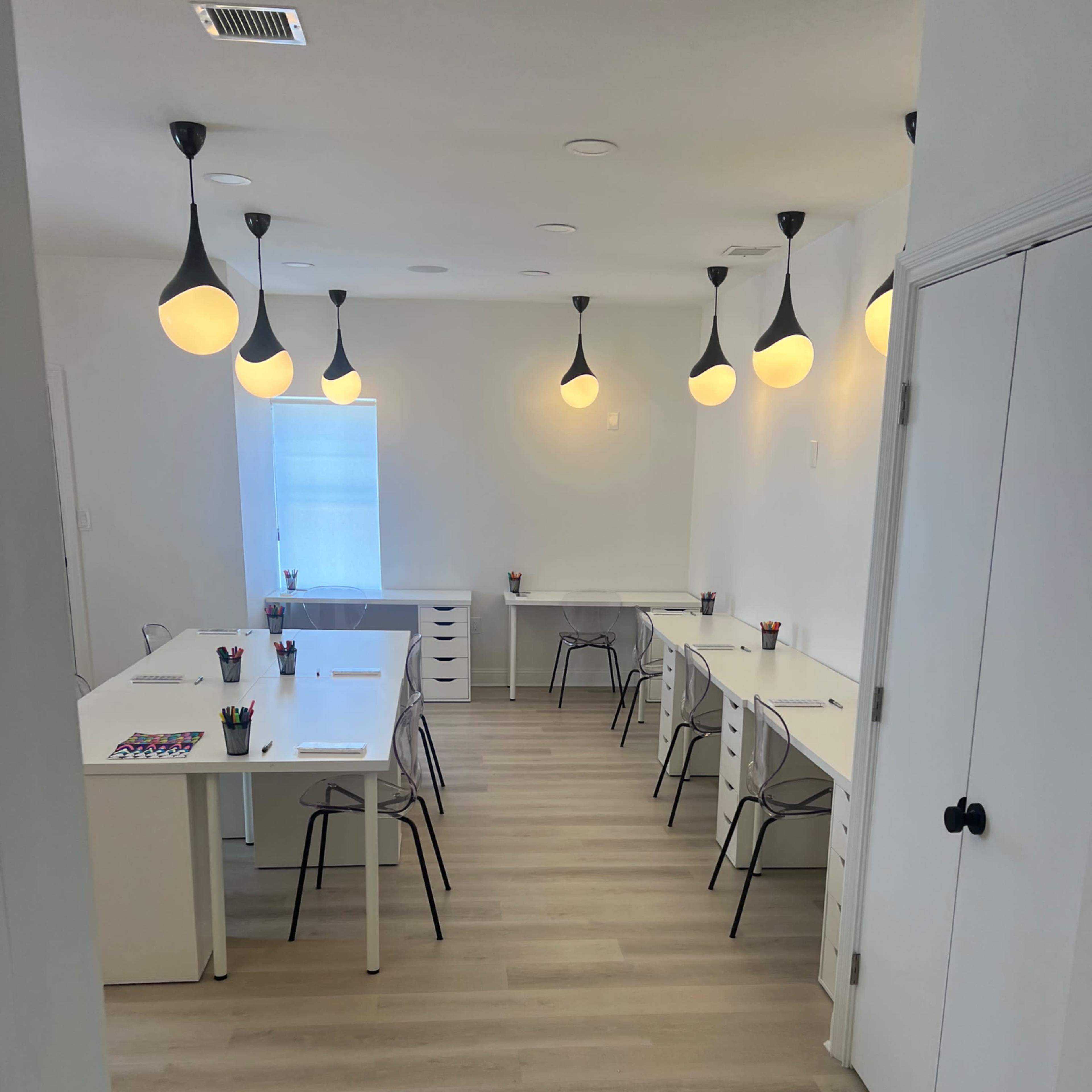 A bright, minimalist workspace with several desks arranged in a spacious room, illuminated by pendant lights.