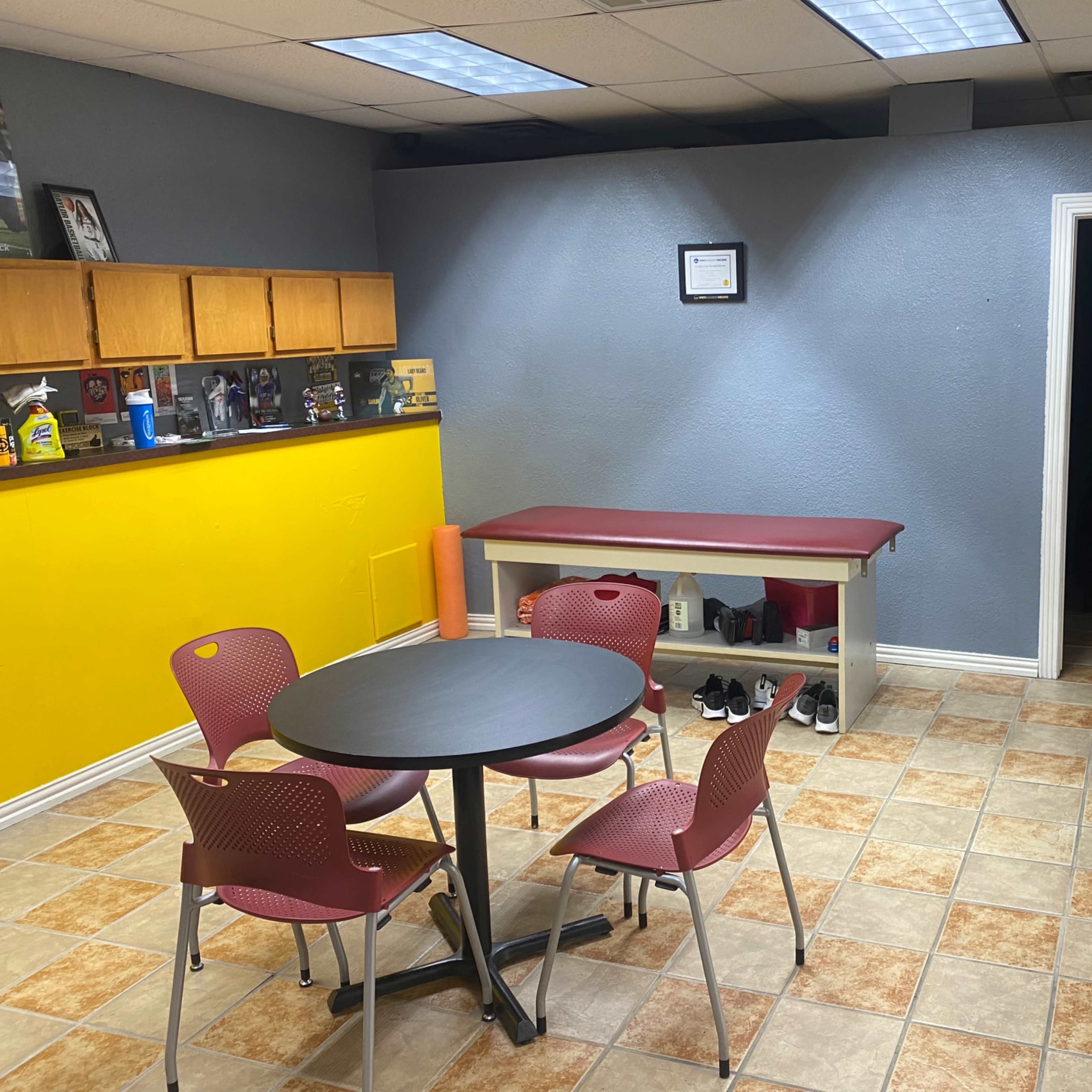 A small waiting area with a round table and four chairs, a treatment table against the wall, and a doorway leading to a darker room.
