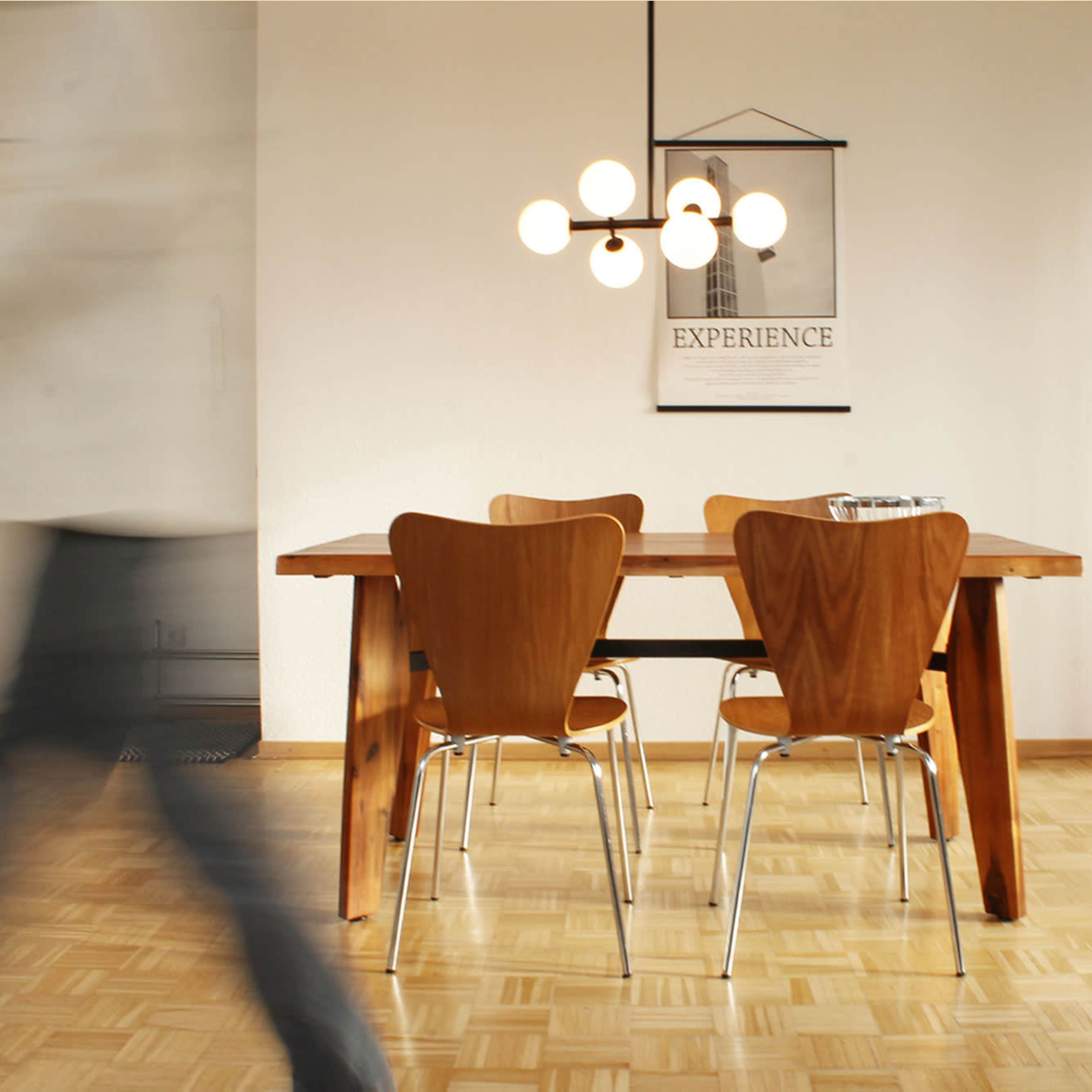 A modern dining area features a wooden table with four chairs and a pendant light hanging above, set against a minimalist white wall.