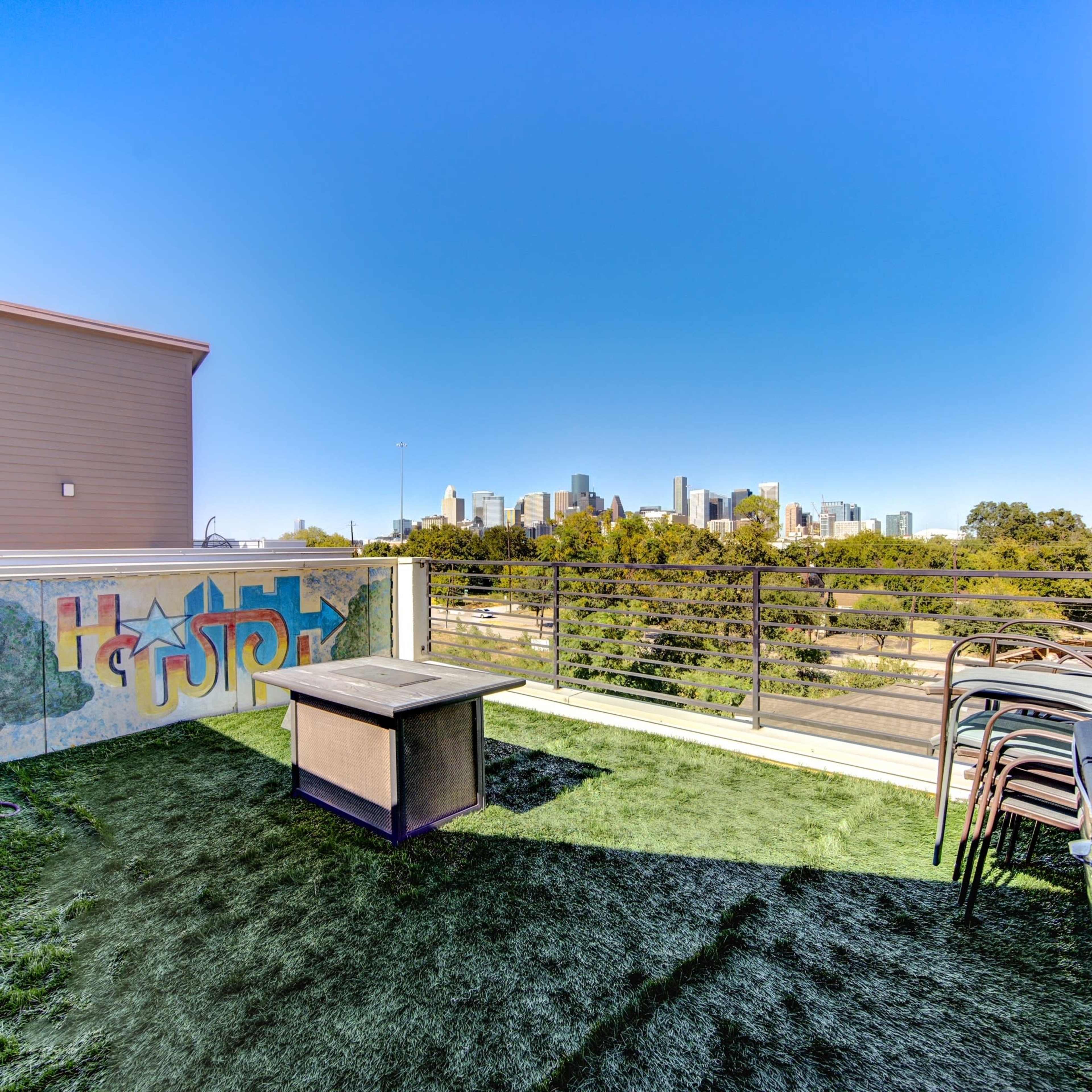 A rooftop terrace features a grassy area, a table, stacked chairs, and a view of a city skyline.