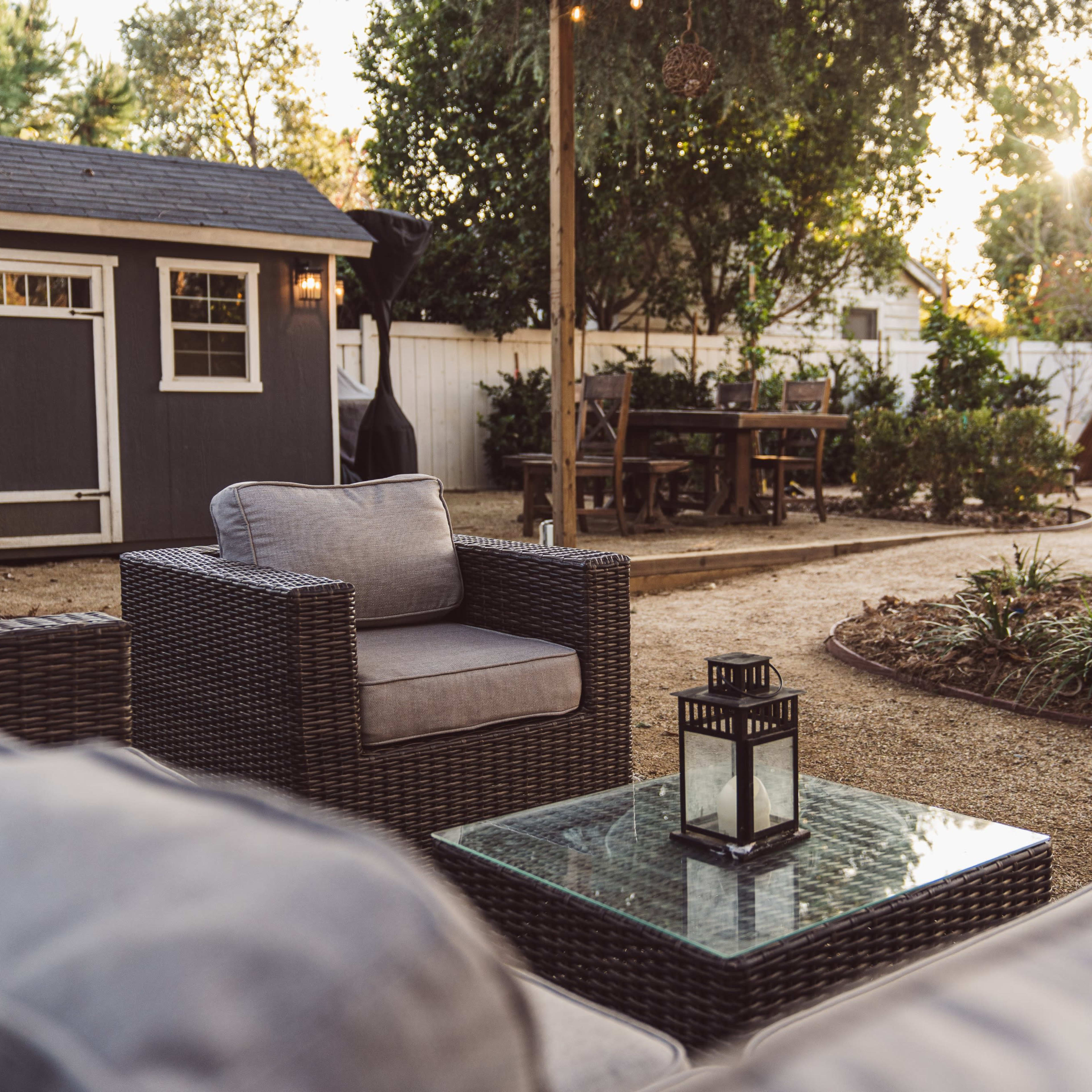 A cozy backyard seating area with wicker chairs, a glass coffee table, and a gray shed in the background, surrounded by greenery and a dining table.