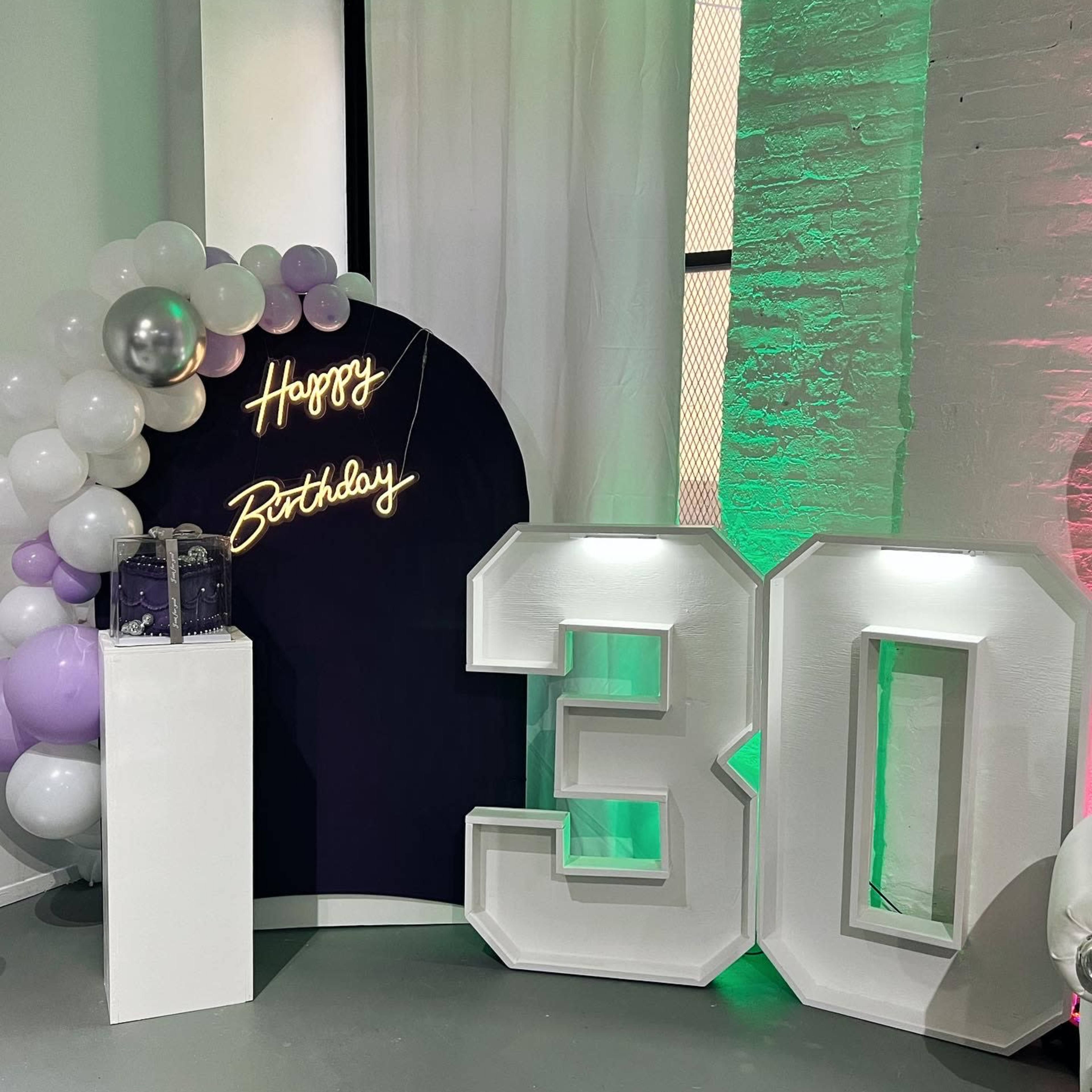 A decorated space for a 30th birthday celebration, with a neon sign that says "Happy Birthday" and large illuminated numbers "30" surrounded by balloons.