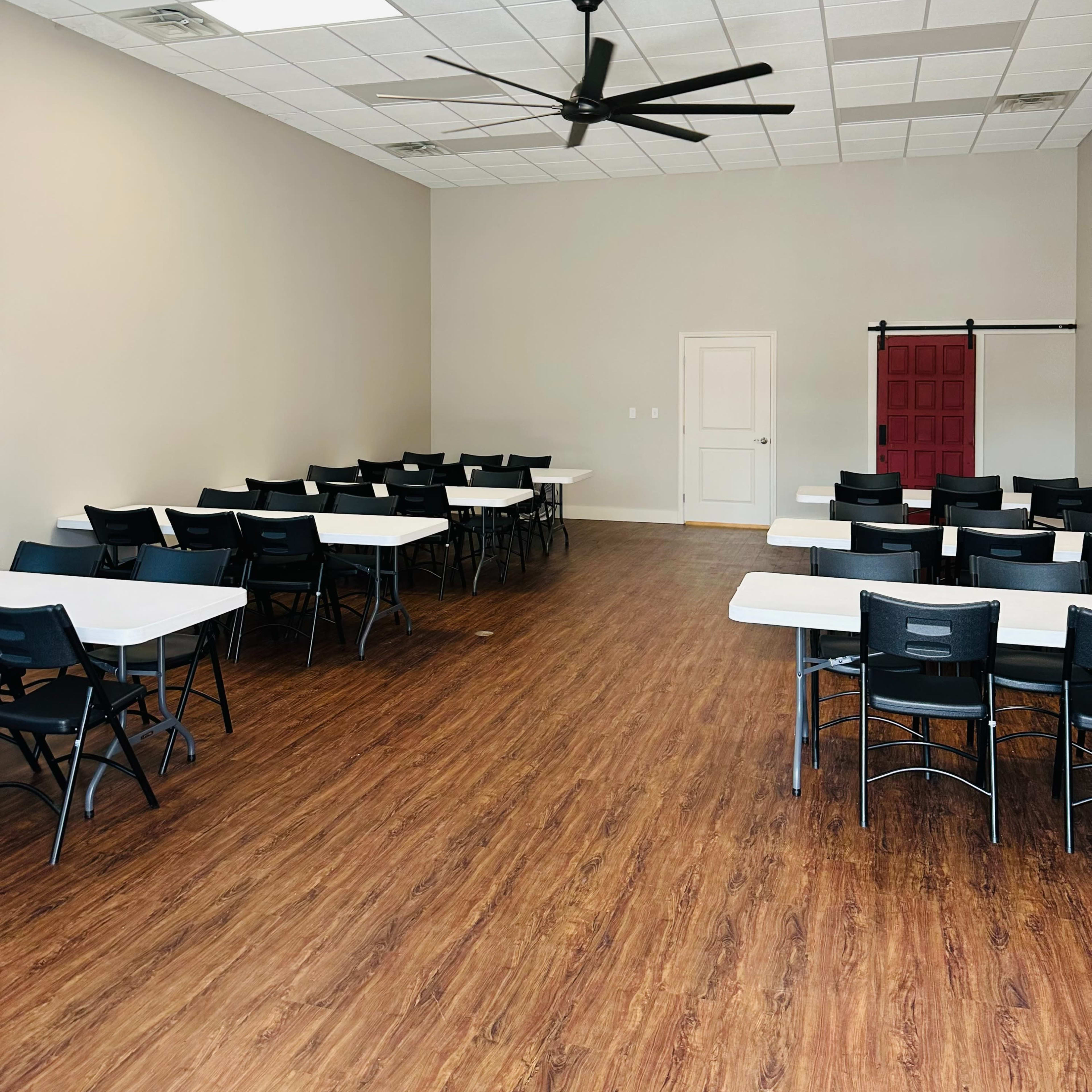 A spacious meeting room with rows of rectangular tables and black chairs, featuring a ceiling fan and a red door at the back.