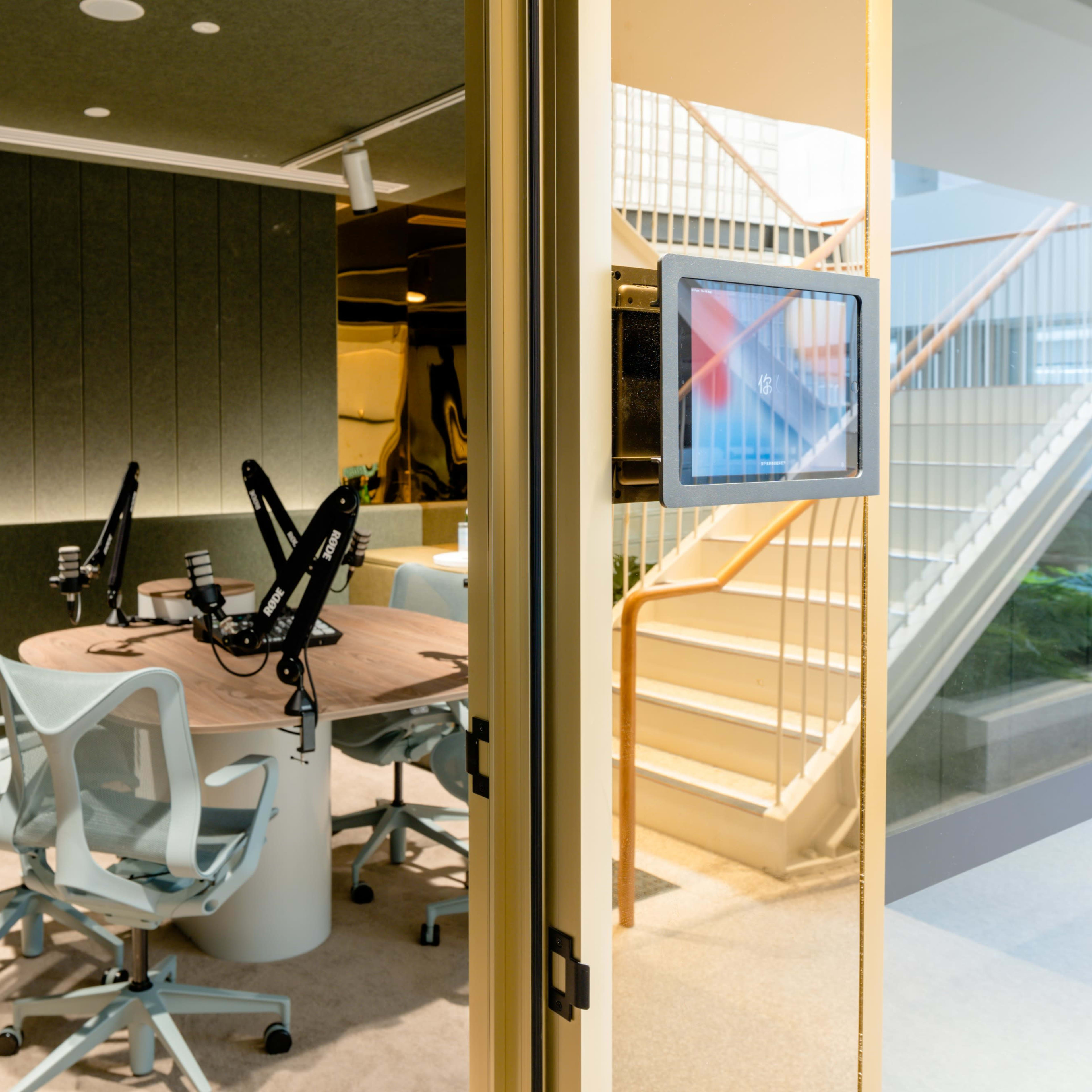 A modern conference room with a round table, four chairs, and two microphones, visible through a glass door with a staircase in the background.