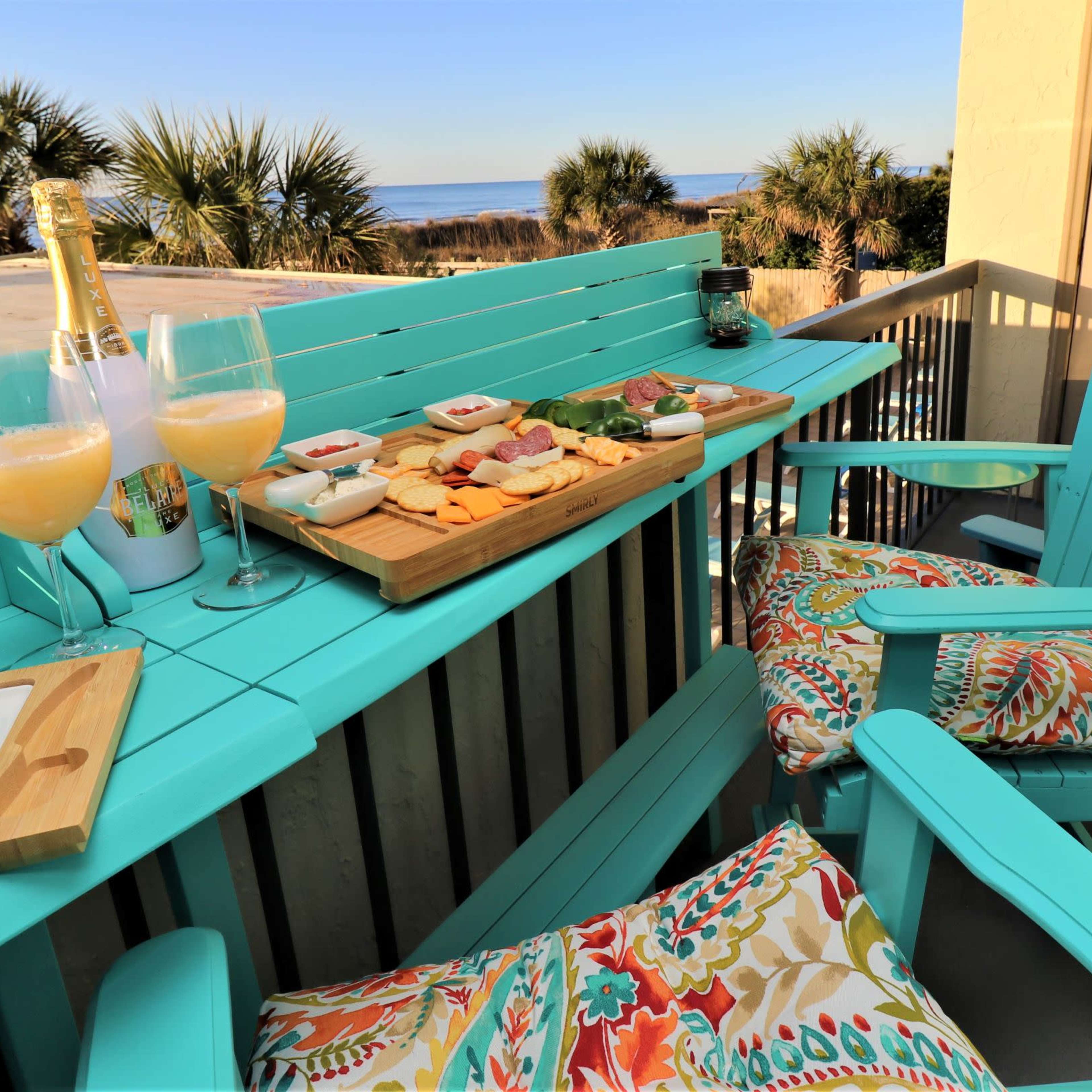 A coastal balcony features a turquoise table set with a cheese platter and drinks, overlooking the ocean and palm trees.