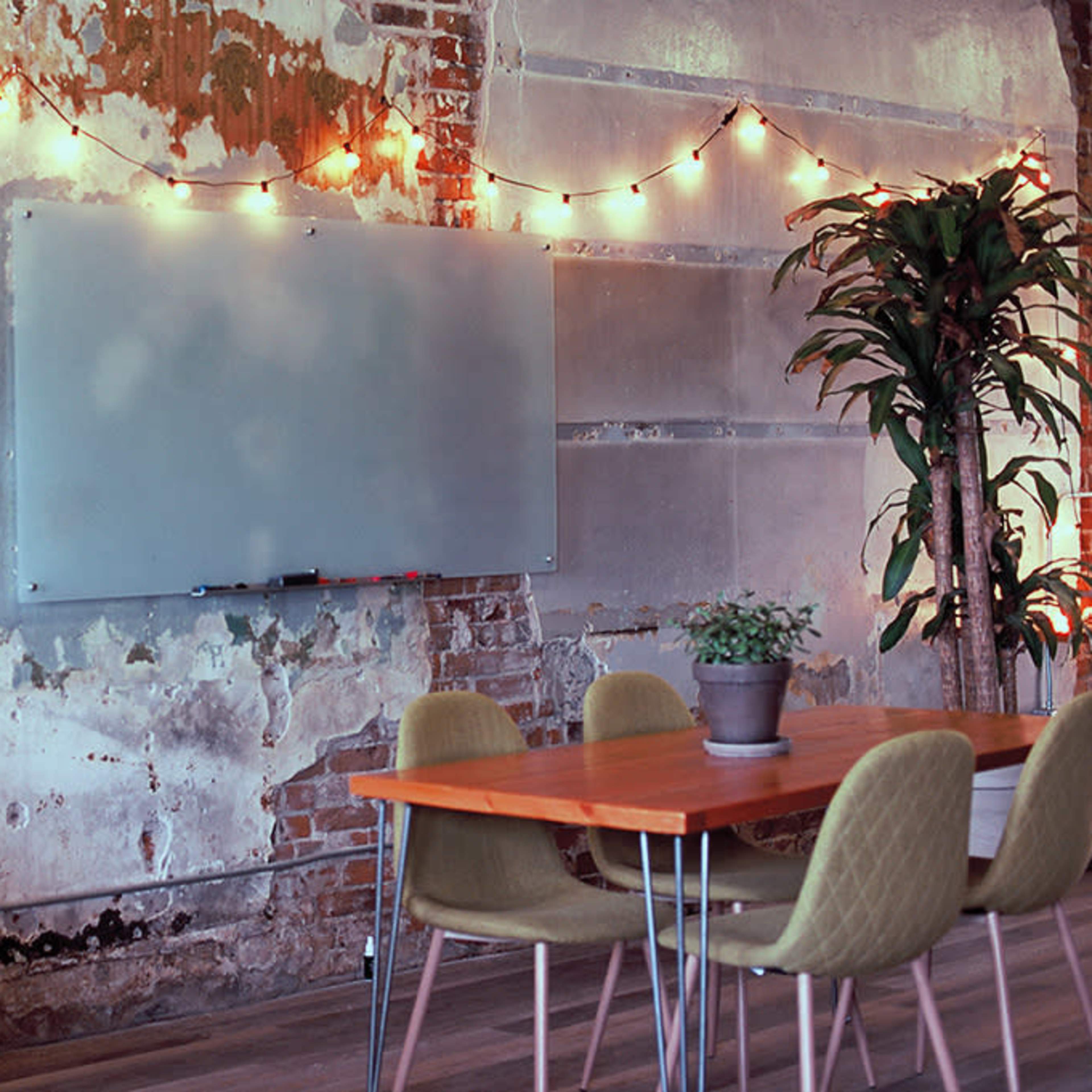 A rustic interior with a weathered wall, string lights, a blank whiteboard, and several tables surrounded by chairs, along with potted plants.
