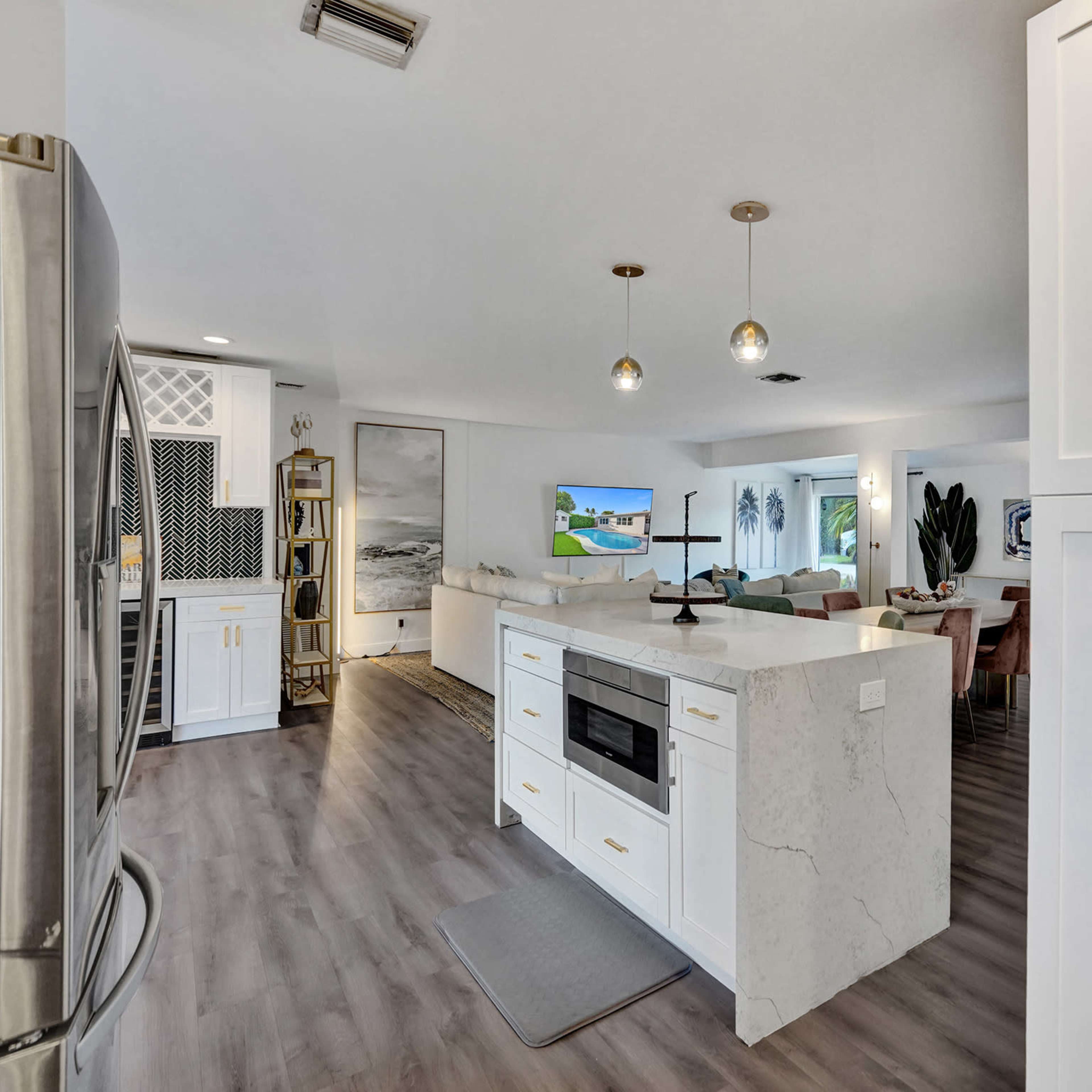 A modern kitchen with white cabinetry, a marble countertop, and an open layout leading to a living area with a television and dining space.