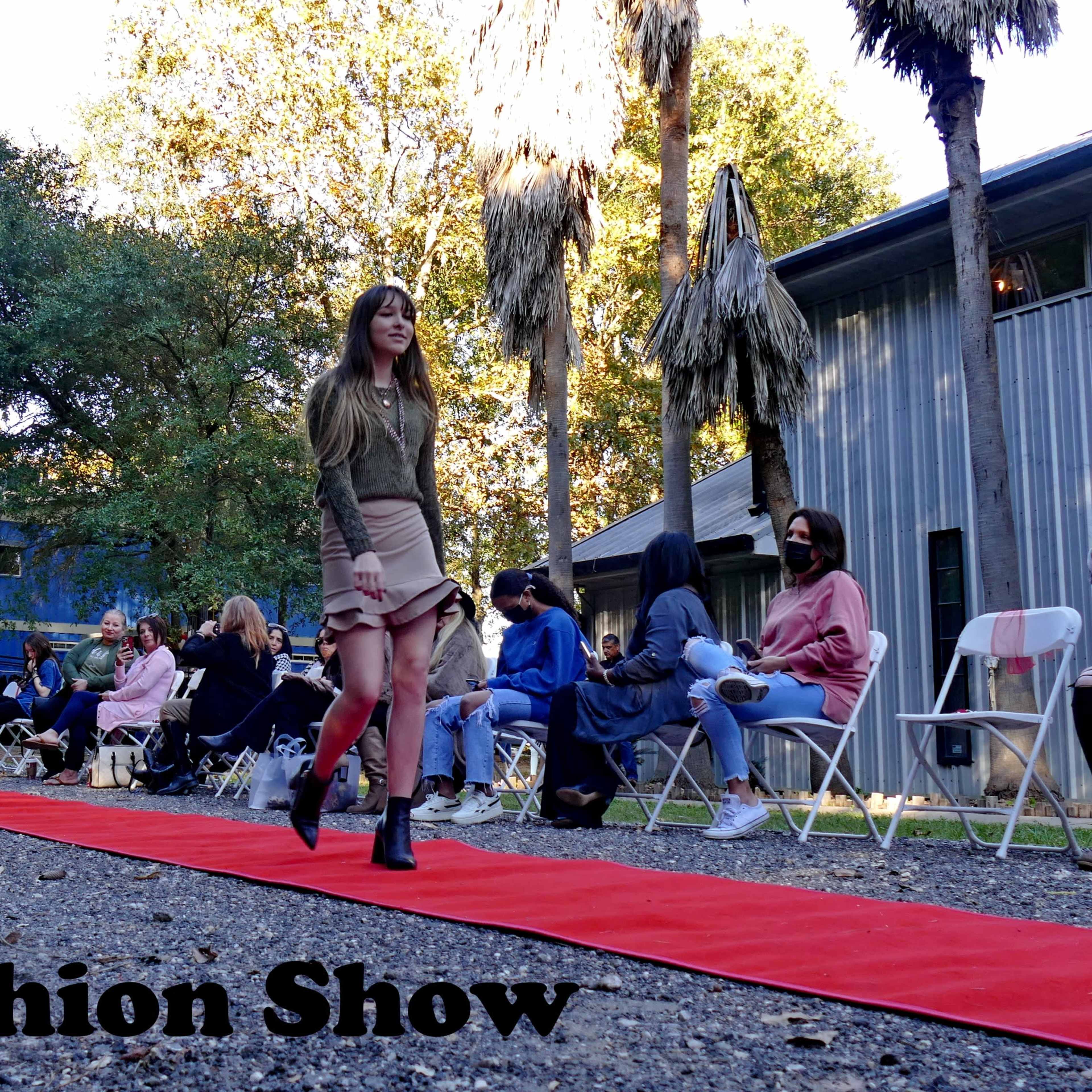 A model walks down a red carpet during a fashion show while an audience sits on either side.