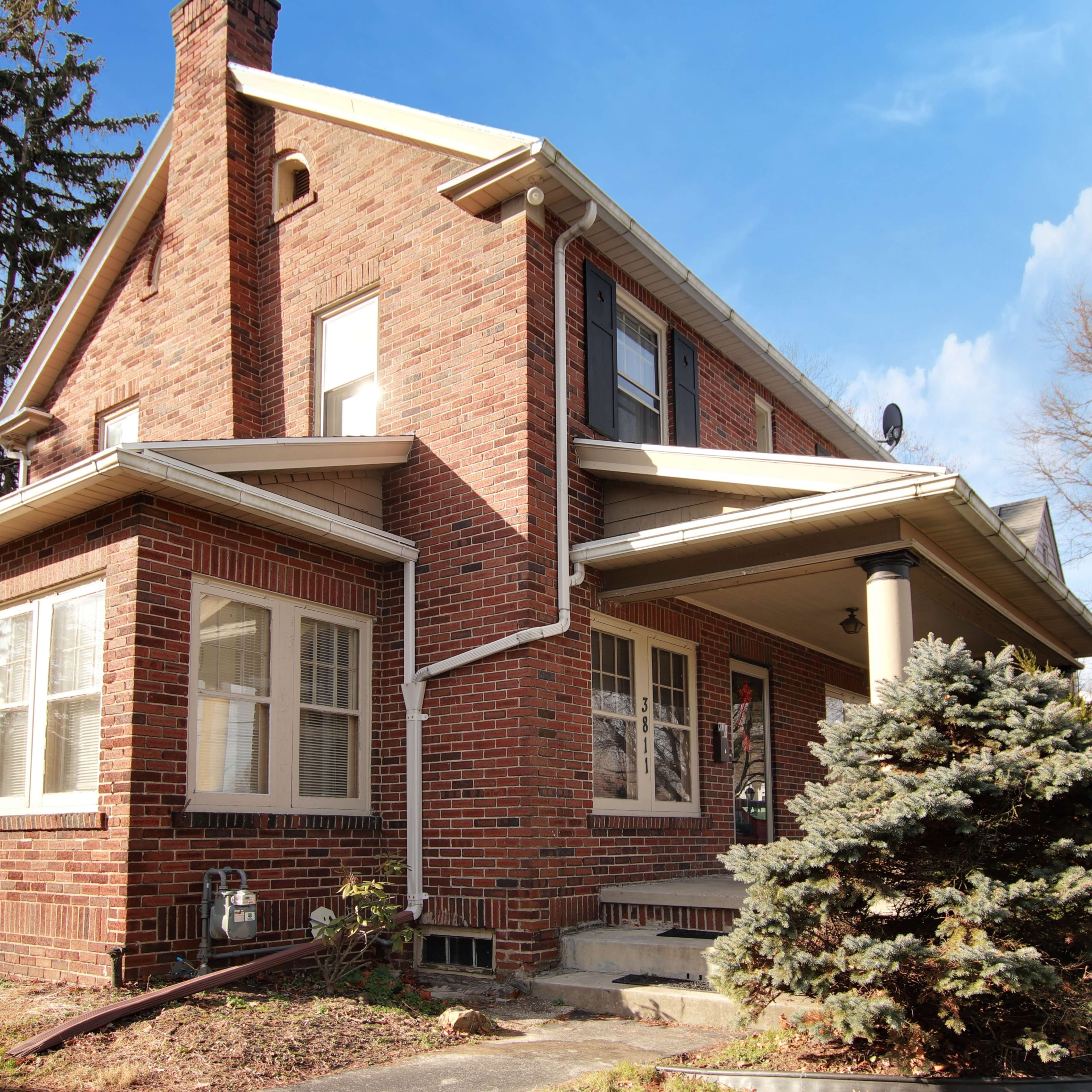 A two-story brick house features a front porch and large windows, surrounded by trees and a landscaped yard.