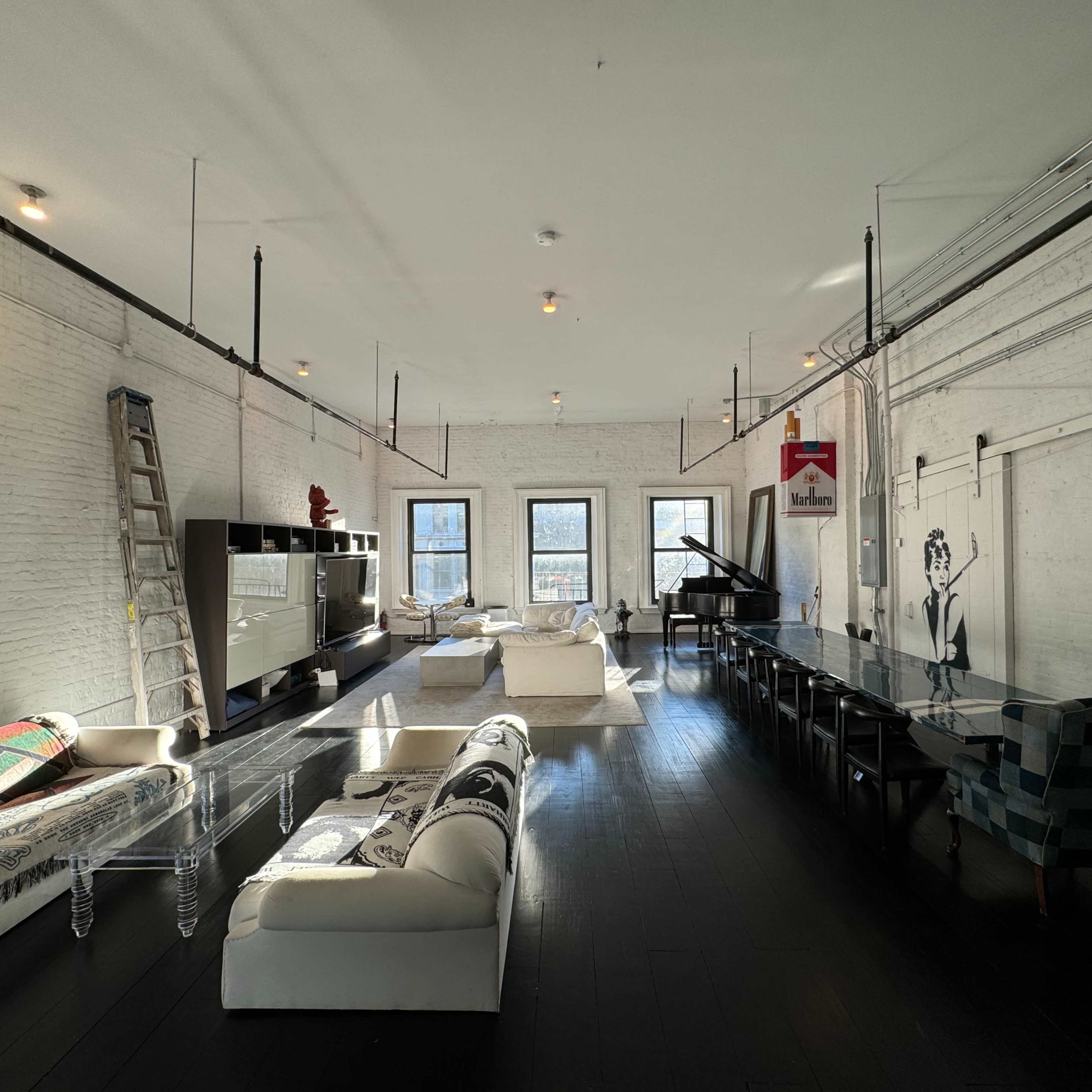 A spacious, modern loft featuring white brick walls, a long dining table, clear acrylic furniture, and a grand piano, with large windows allowing natural light to fill the space.