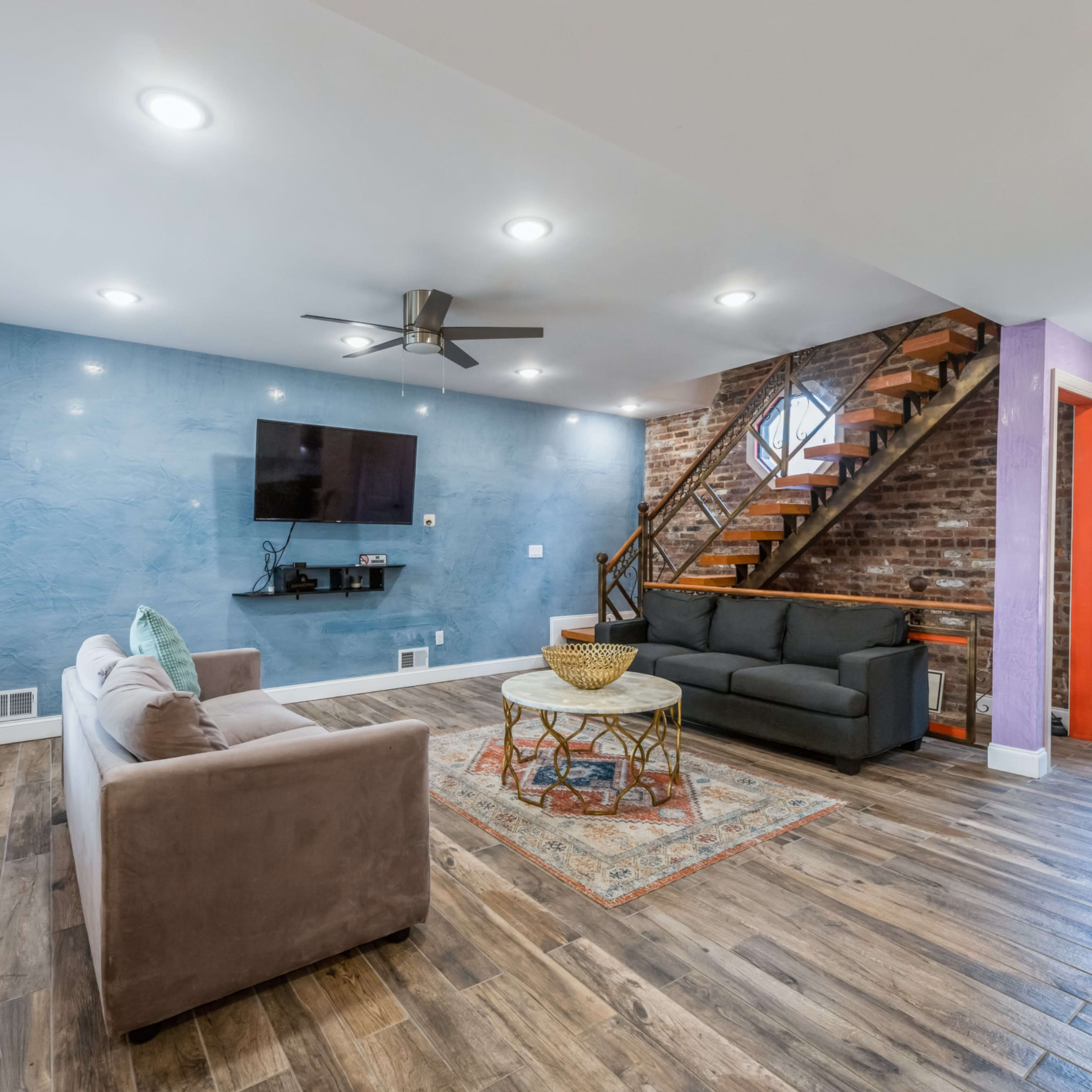 A living room with a blue accent wall, two sofas, a round coffee table, a television mounted on the wall, and a staircase leading to an upper level.