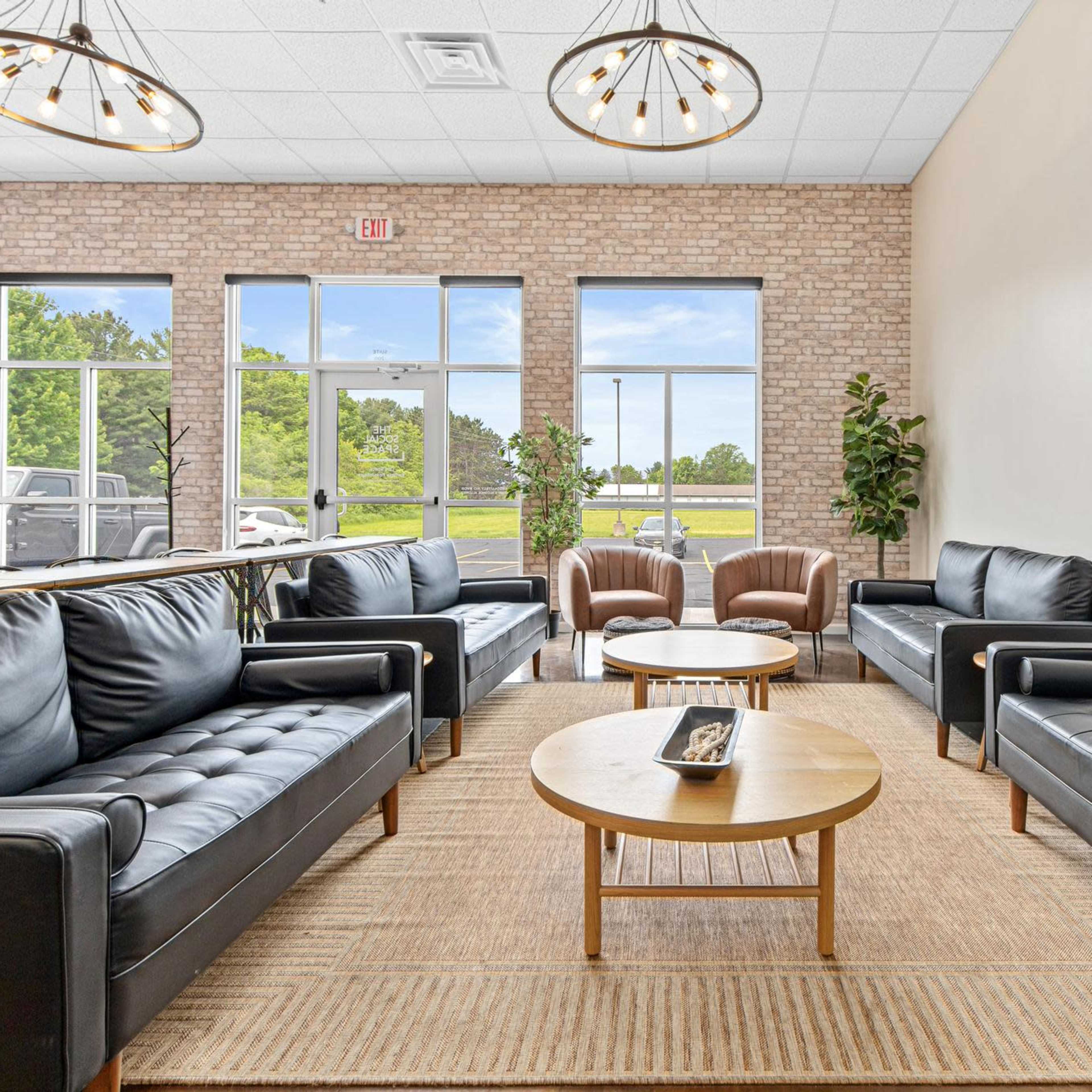 A modern seating area with four black leather sofas, two brown armchairs, and a round wooden coffee table, set against a backdrop of large windows and a brick wall.