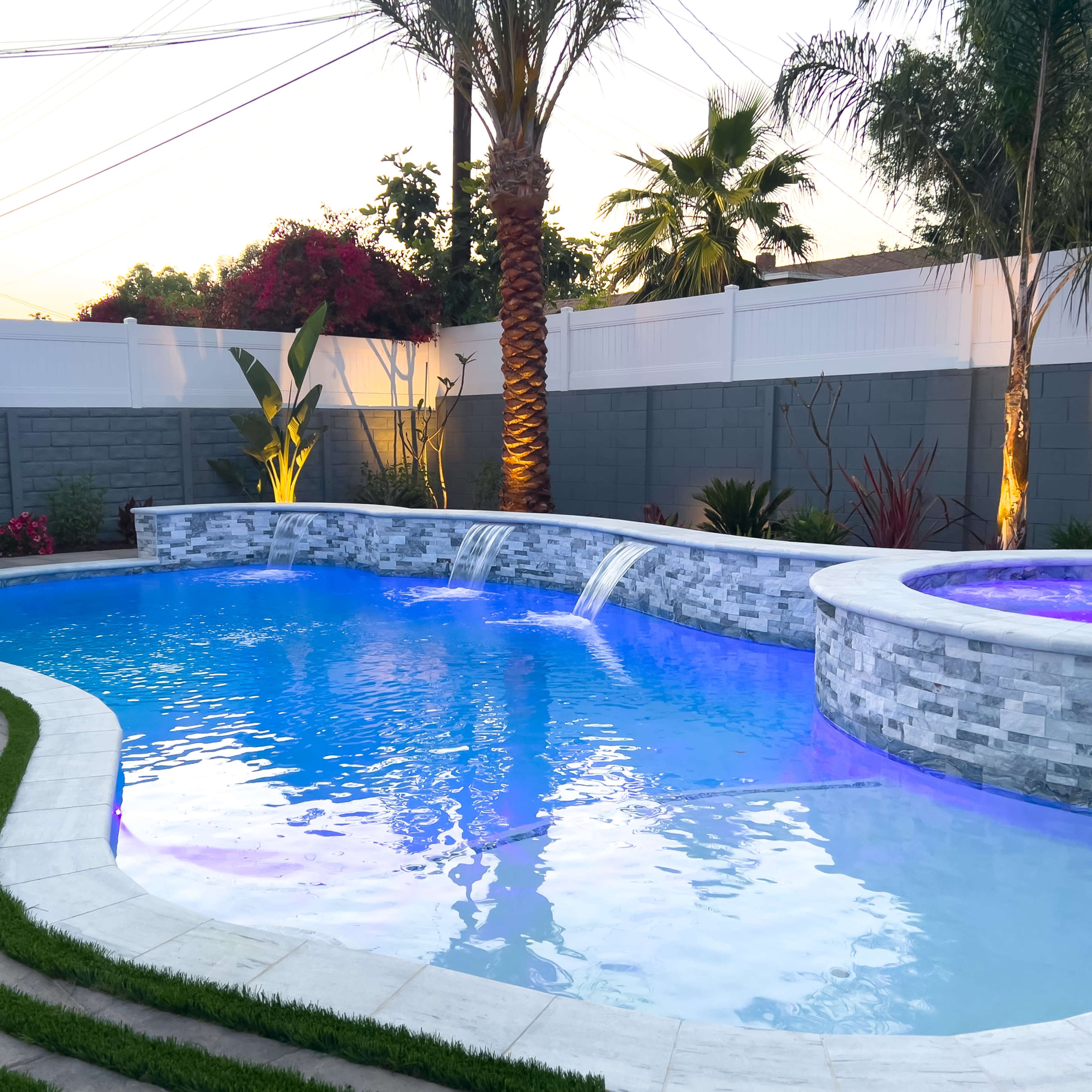 A backyard swimming pool featuring a stone waterfall and a spa, surrounded by landscaped greenery and illuminated at dusk.