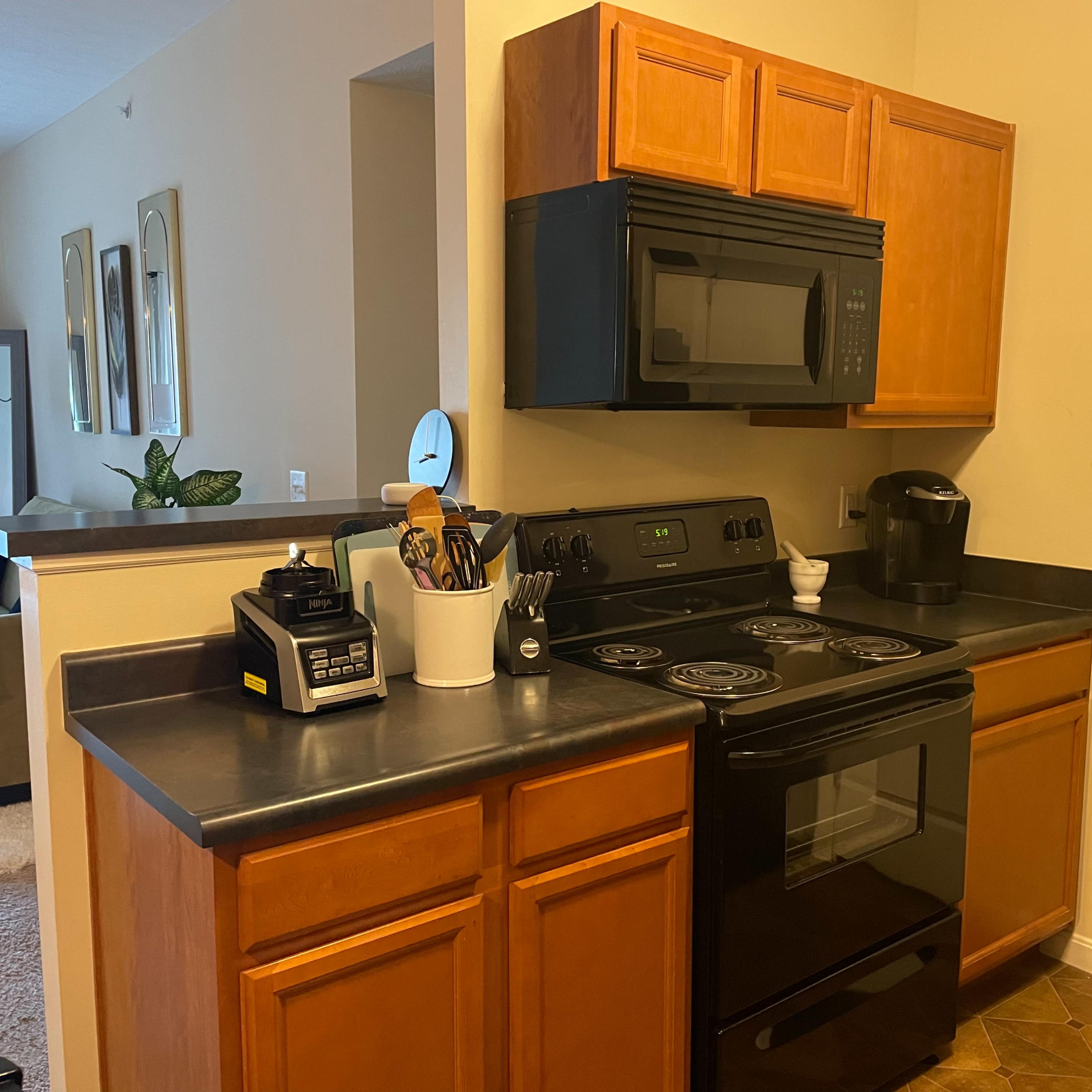 A kitchen area featuring wooden cabinets, a black microwave and stove, and various kitchen appliances on the countertop.