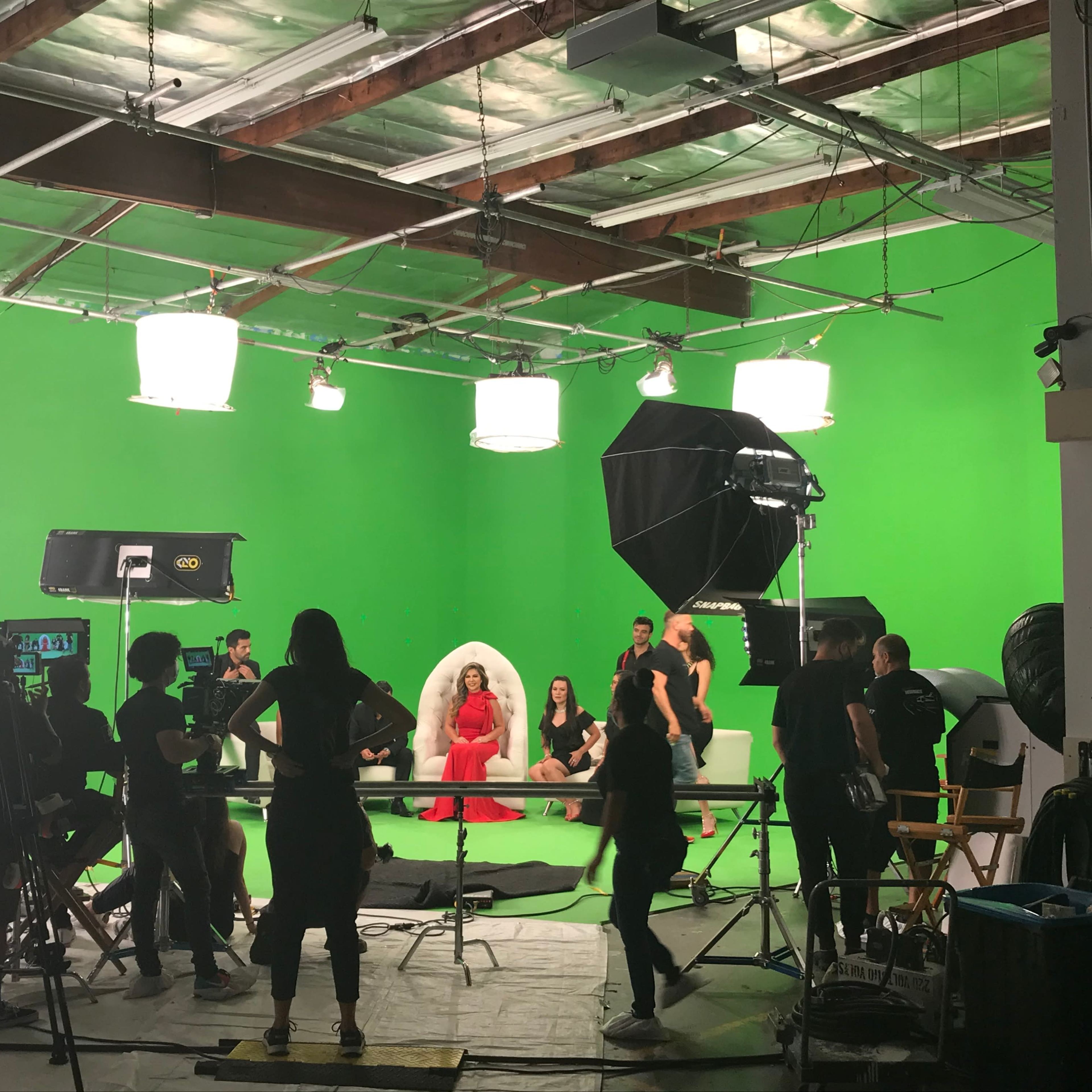 A film crew sets up in a green screen studio, featuring a central figure in a red dress surrounded by additional people and various lighting equipment.