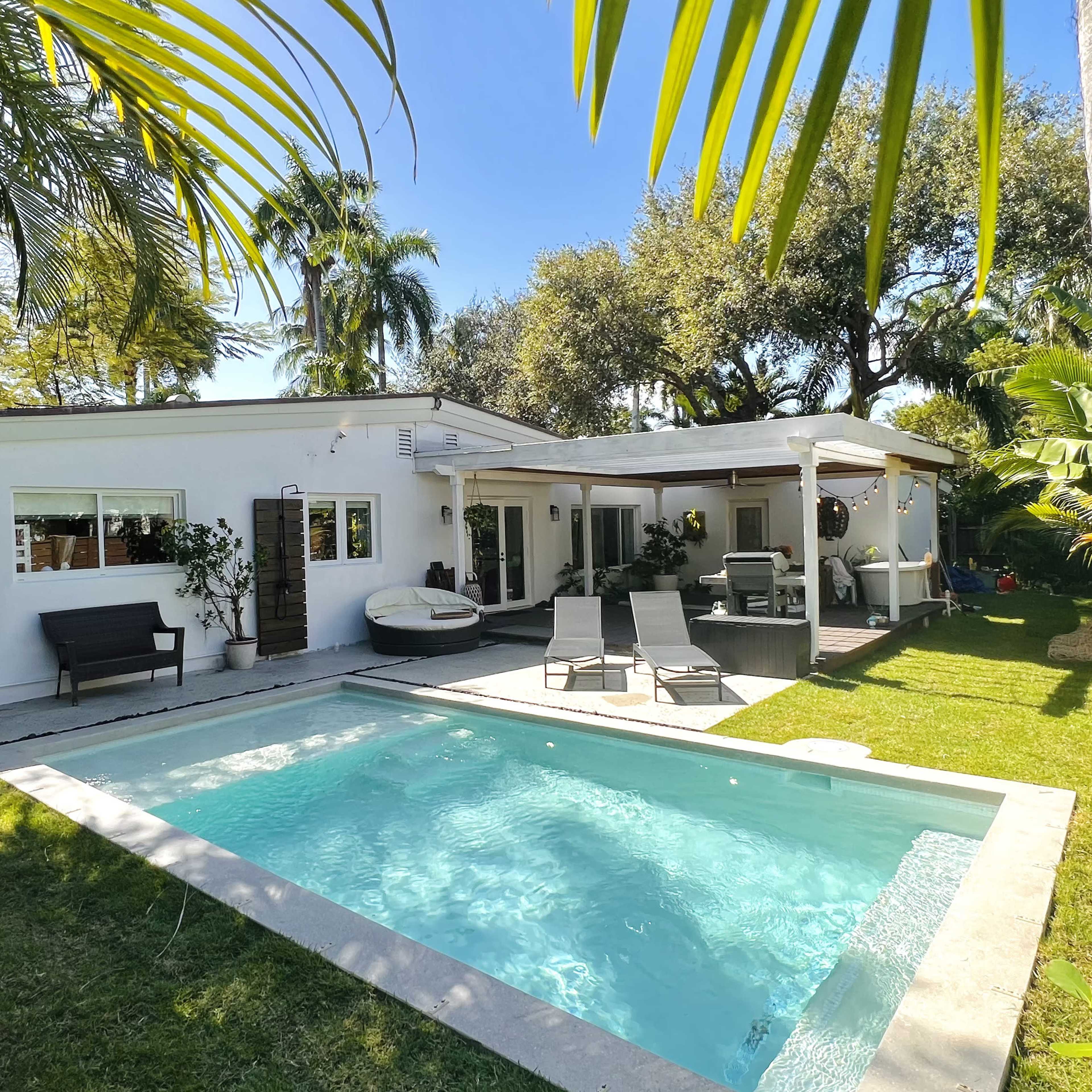 A small, modern house with a pool and patio area is surrounded by lush greenery and palm trees.