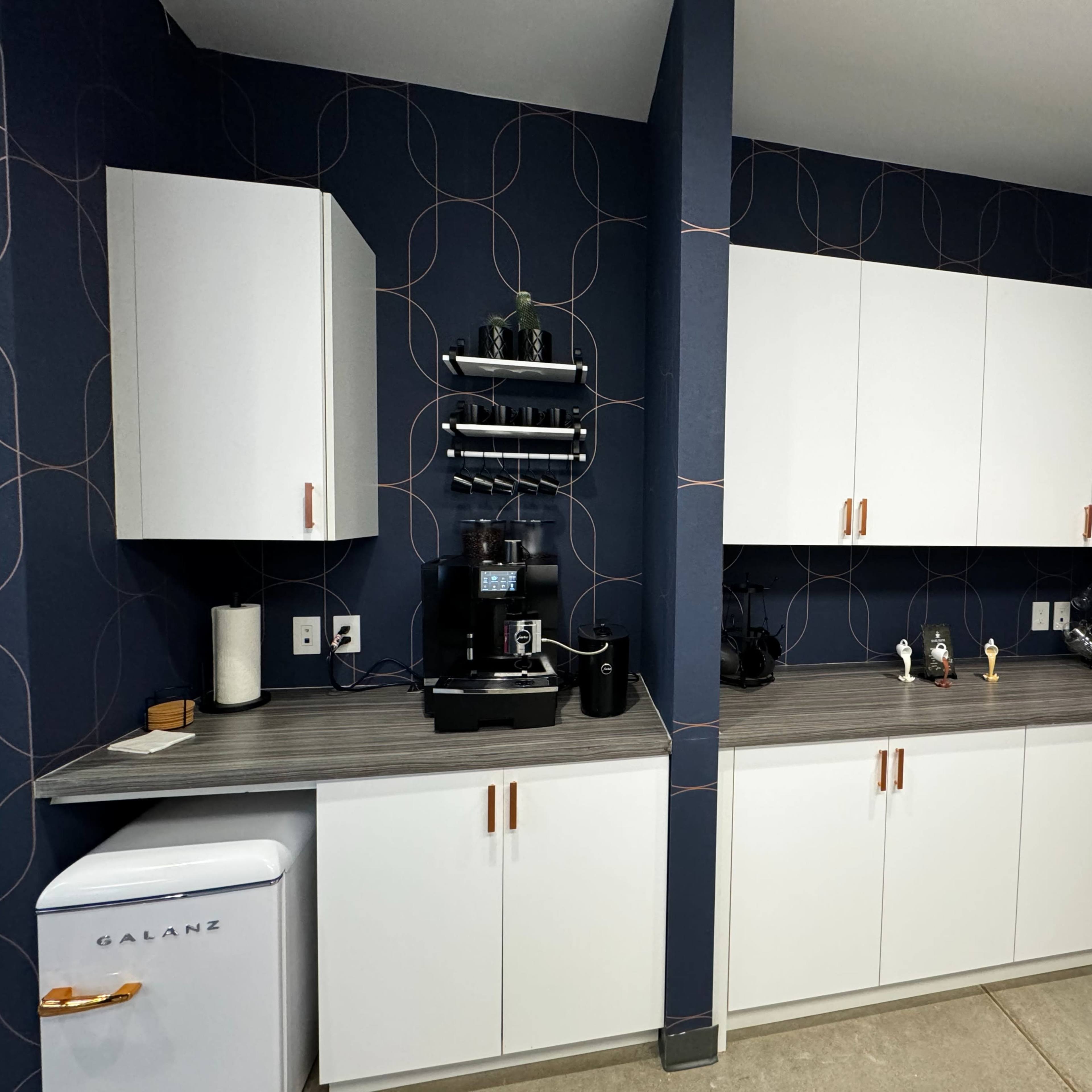 A modern kitchen area featuring a dark blue patterned wall, white cabinets, a countertop with a coffee machine, and a small refrigerator.