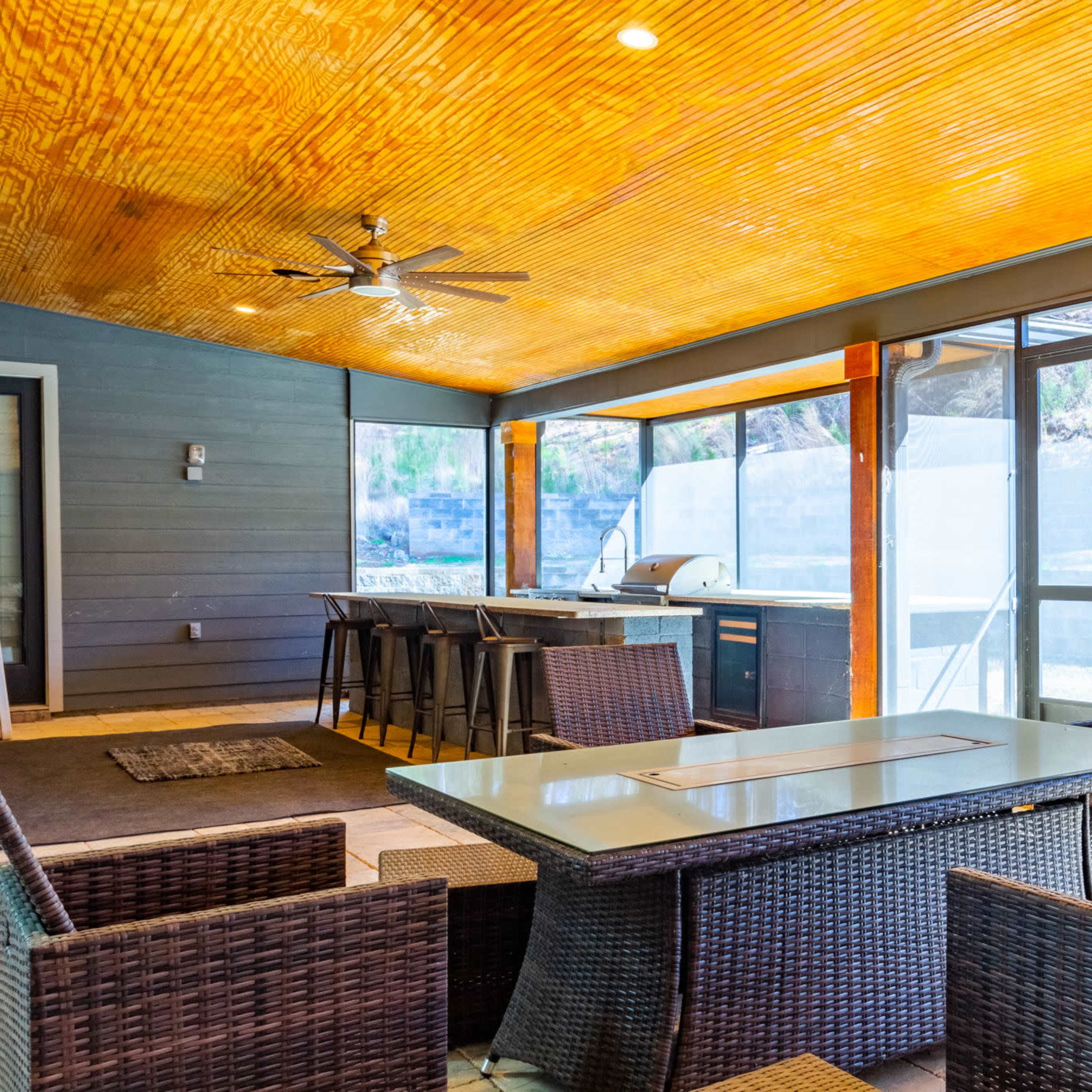 A spacious outdoor patio featuring a seating area with wicker chairs and a glass table, adjacent to a kitchen space with a grill and counter.