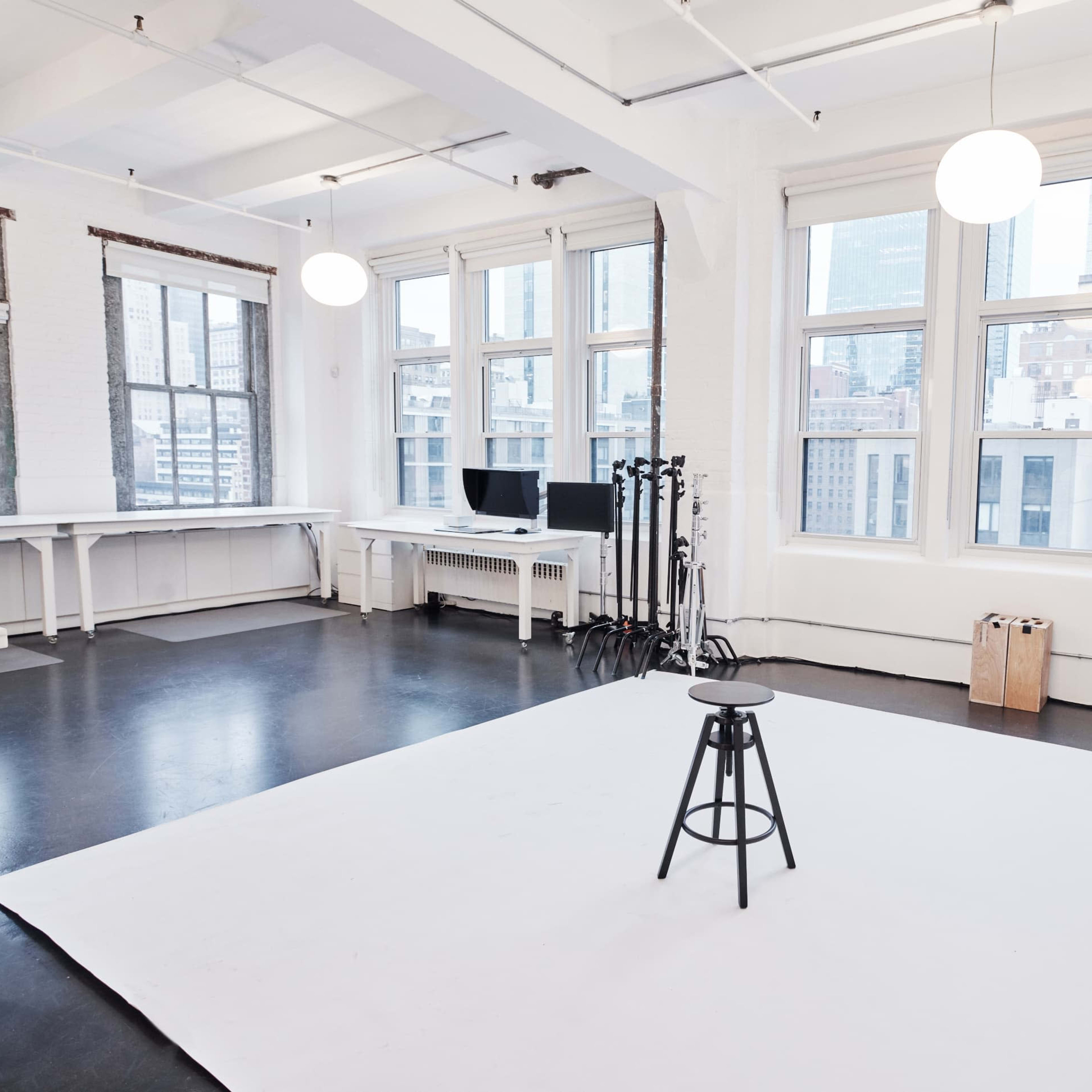 A spacious photography studio with large windows, a white backdrop, a black stool, and studio lighting equipment.